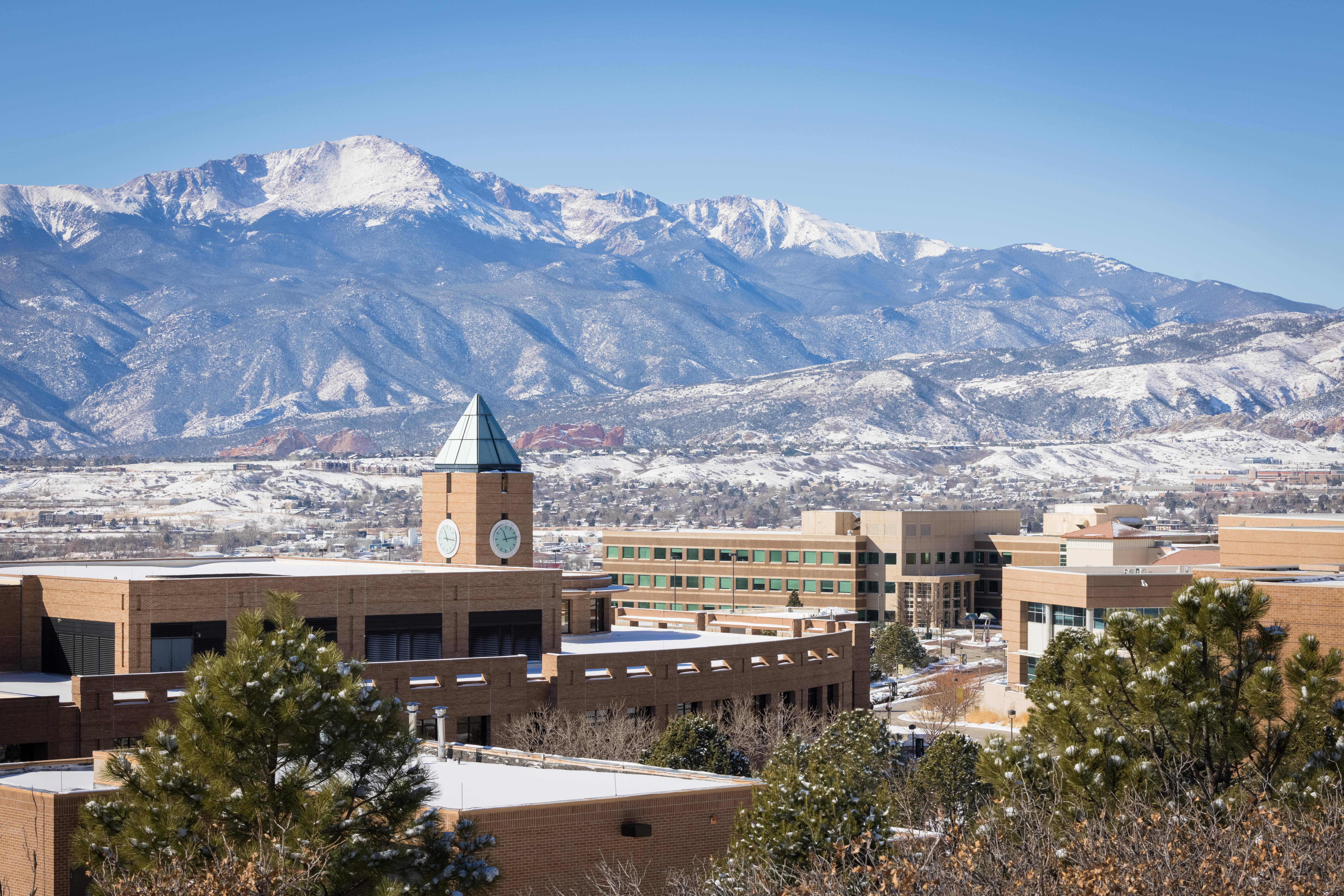 科羅拉多大學雪山環抱的美麗校園 壁紙 — 白雪覆蓋的山脈在現代磚砌建築後方升起，湛藍的天空下，前景有常綠樹木。