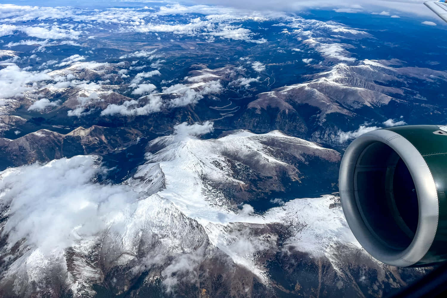 科羅拉多山脈的壯麗風景 壁紙 — 從飛機窗戶俯瞰，前景是噴射引擎，下方是積雪的山脈和雲層。