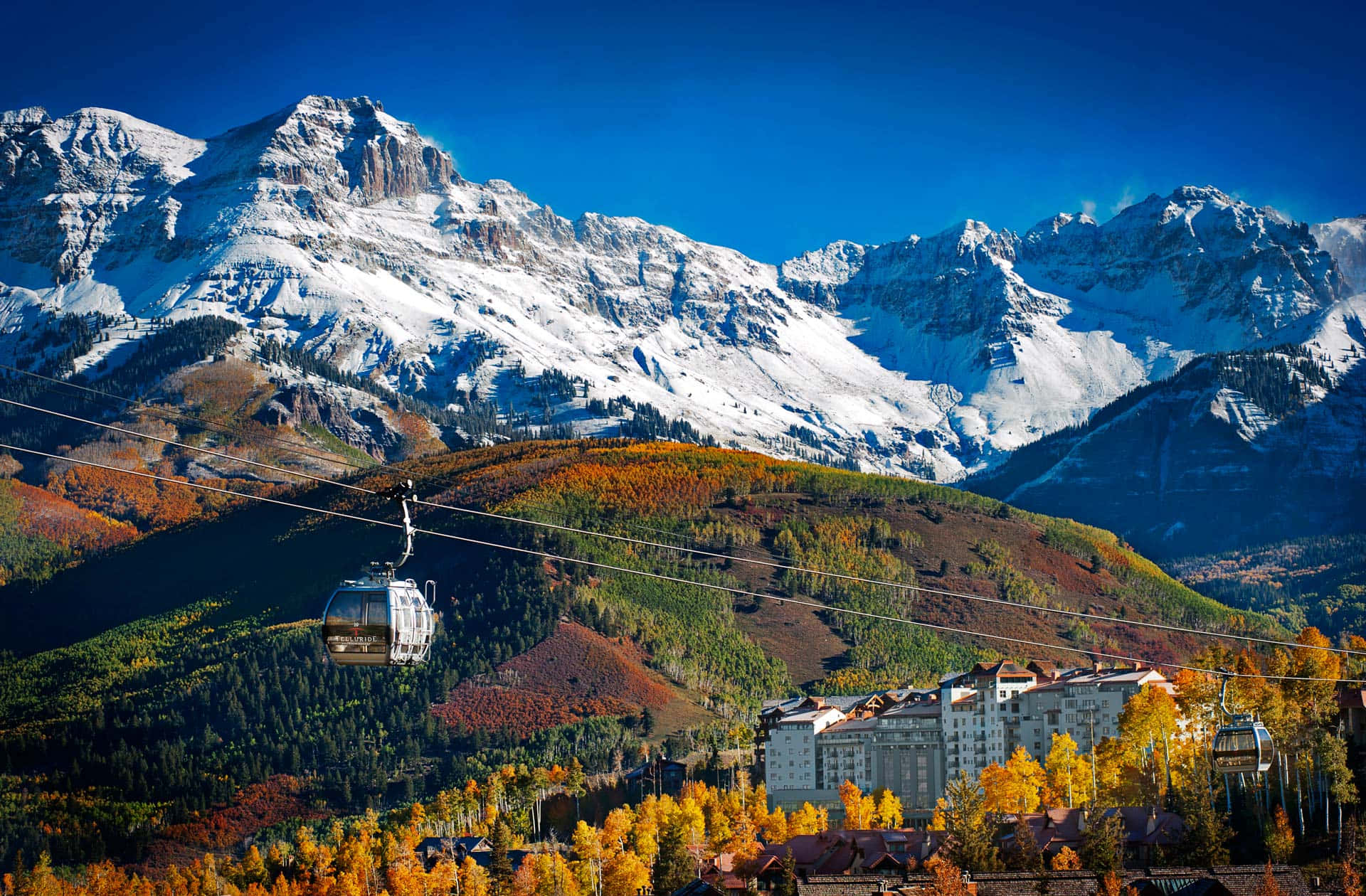 科羅拉多山脈的壯麗風景 壁紙 — 一架纜車在晴朗藍天下，滑過一個充滿活力的秋季山谷，山谷中有積雪山峰和一個度假小鎮。