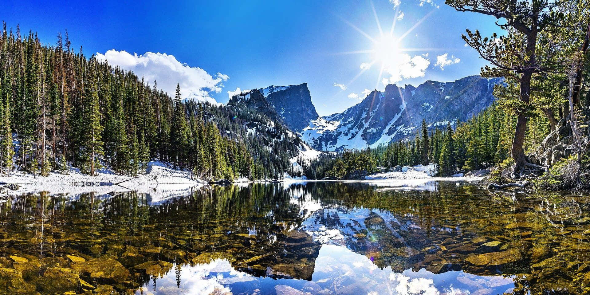 科羅拉多山脈的如畫美景 壁紙 — 燦爛的陽光照耀在積雪的山峰上，完美地倒映在寧靜的高山湖泊中，周圍是常綠森林。