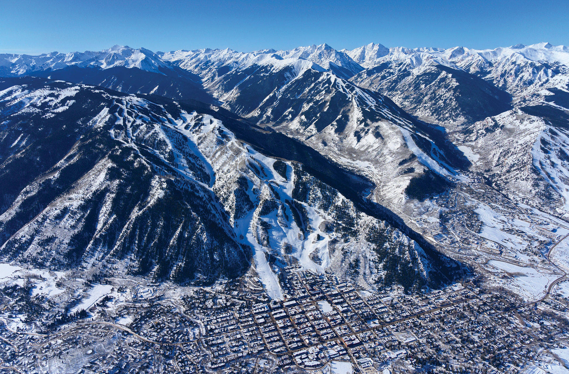 科羅拉多州阿斯彭空中攝影 壁紙 — 白雪覆蓋的山峰鳥瞰圖，滑雪道在清澈藍天下延伸至冰封的山谷小鎮。