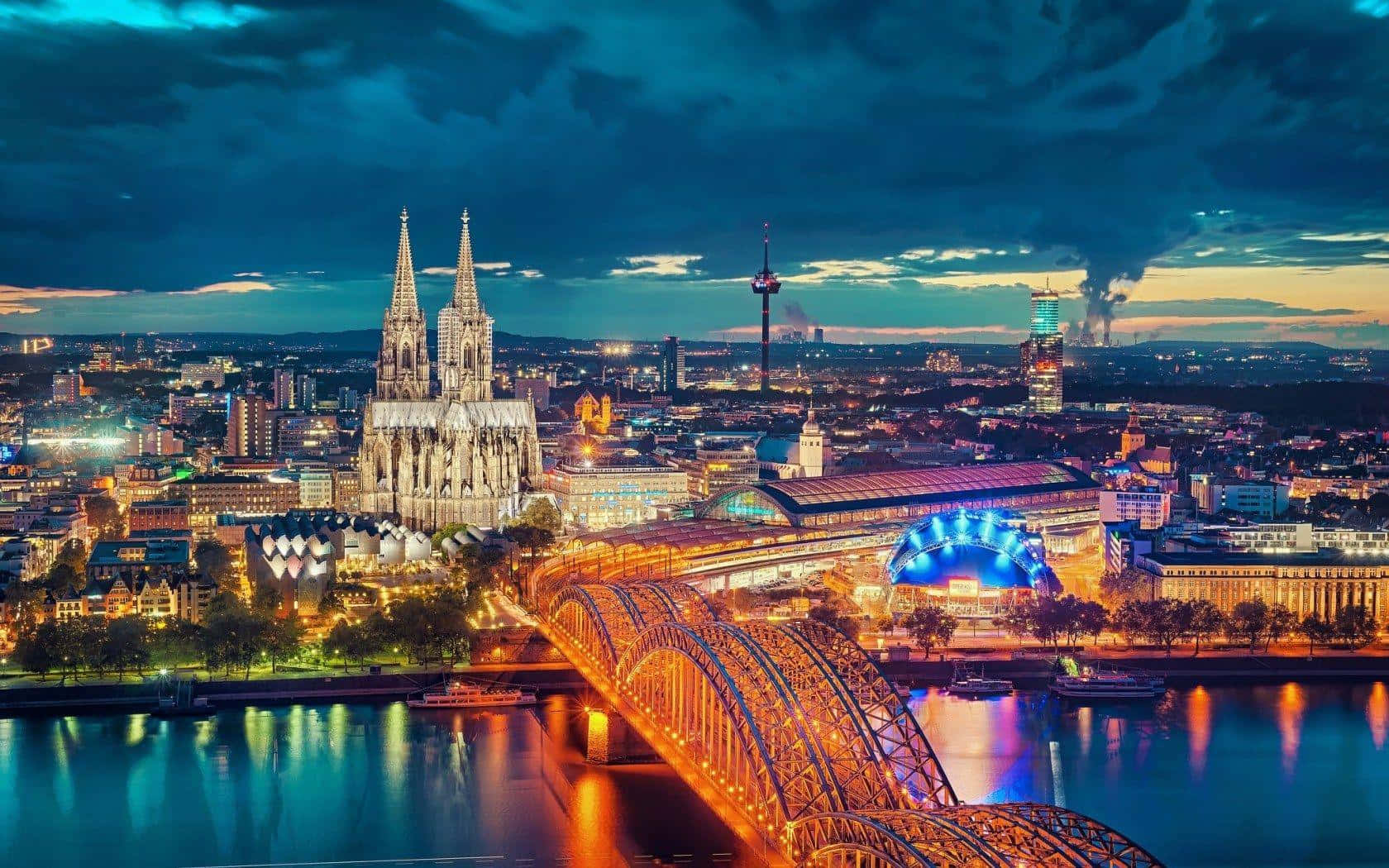 Germany 桌布 壁紙 — 科隆大教堂和霍亨索倫橋在萊茵河上暮色天空下閃耀，城市燈光倒映在水中。