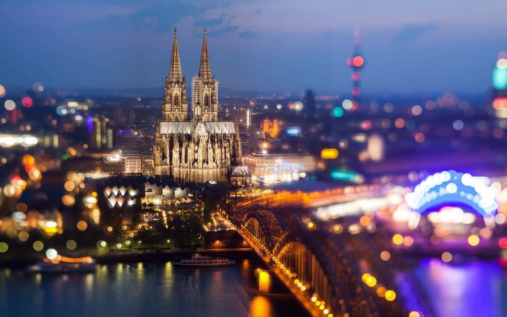 Germany 桌布 壁紙 — 科隆大教堂在暮色天空下被照亮，周圍模糊的城市燈光和河流倒影閃爍著光芒。