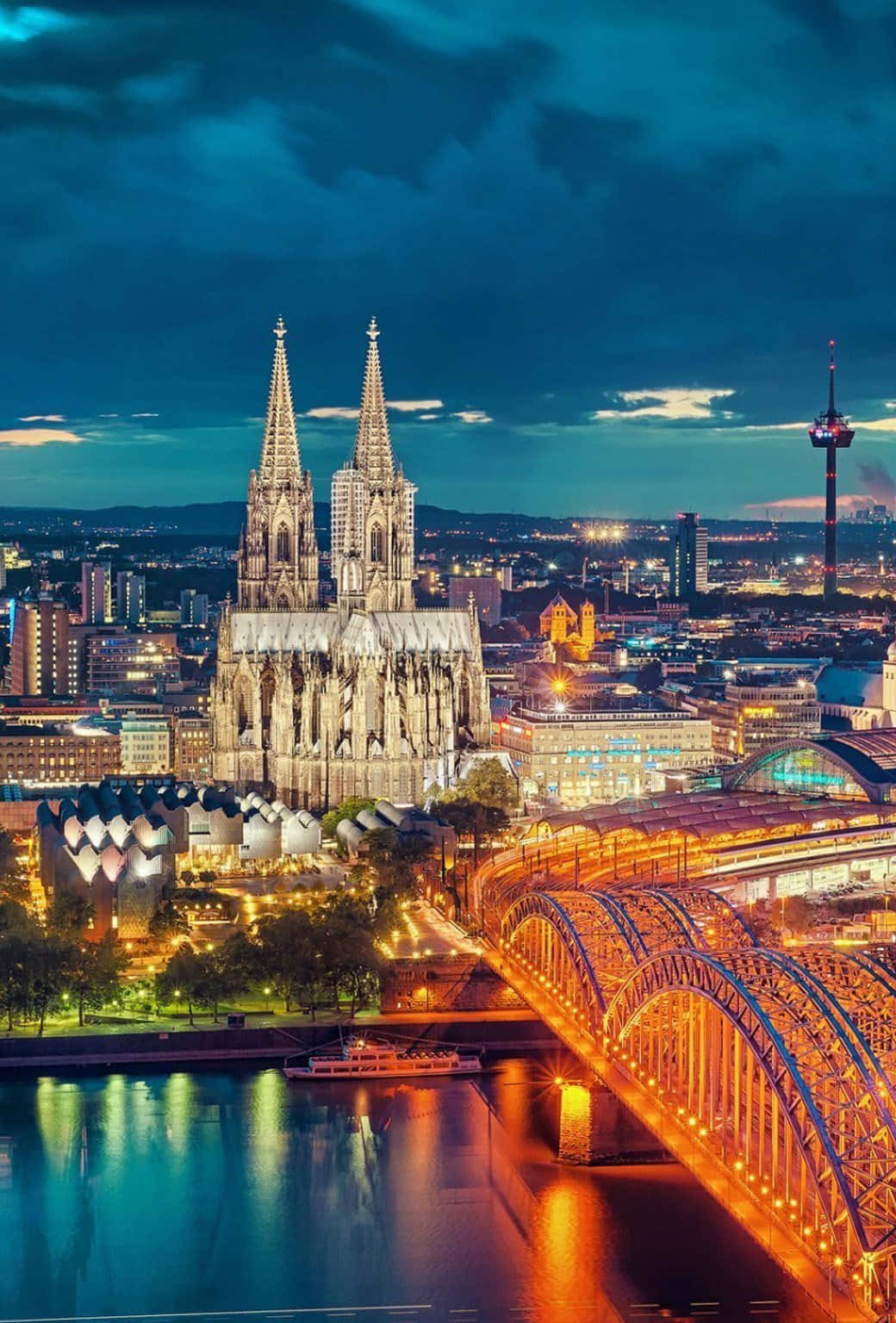 Germany 桌布 壁紙 — 科隆大教堂在黃昏時被照亮，聳立在閃爍的城市景觀之上，下方河流反射著一座亮燈的橋樑。