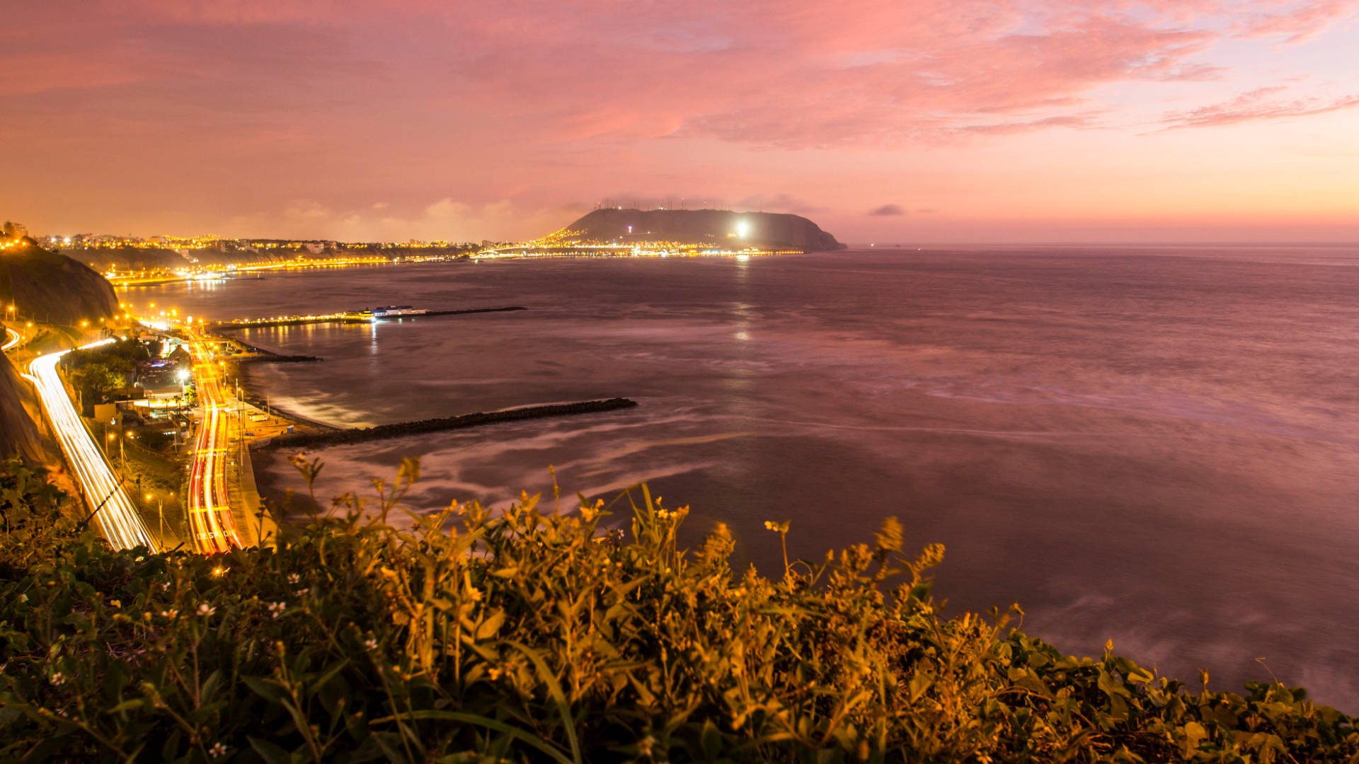 秘魯利馬海岸夜景 壁紙 — 充滿活力的黃昏海岸場景，沿著彎曲的高速公路閃爍的城市燈光，以及在粉紅色和橙色天空下的遠山。