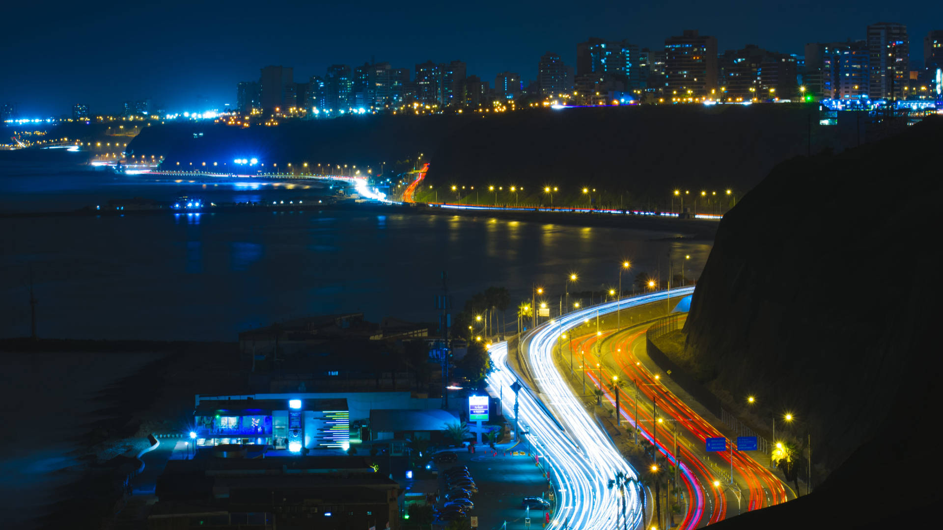 秘魯科斯塔維爾德夜景 壁紙 — 在黑暗的天空下，汽車沿著河岸蜿蜒行駛，留下發光的燈光軌跡，構成充滿活力的夜景。