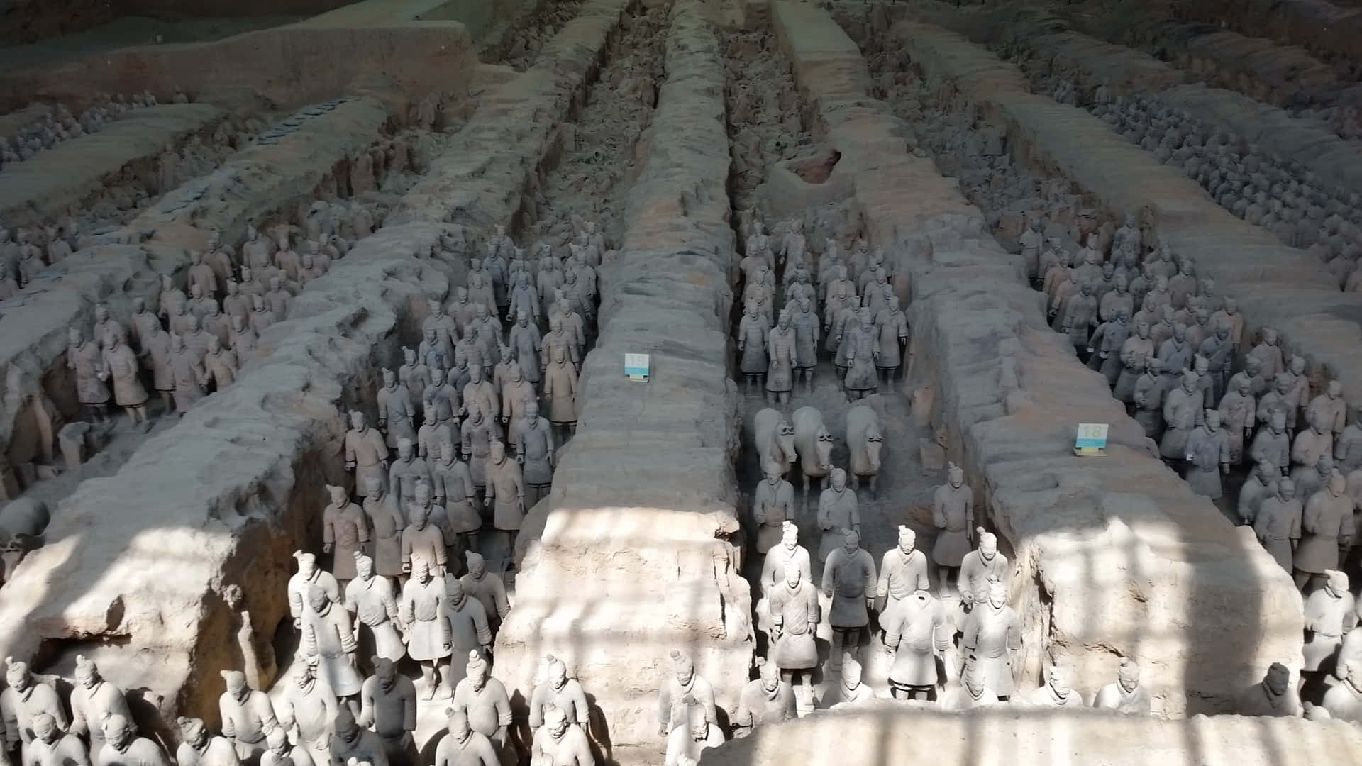 秦始皇陶俑軍隊巨像 壁紙 — 一排排古老的兵馬俑整齊地排列在泥土掩埋的坑內，沐浴在自然光中。
