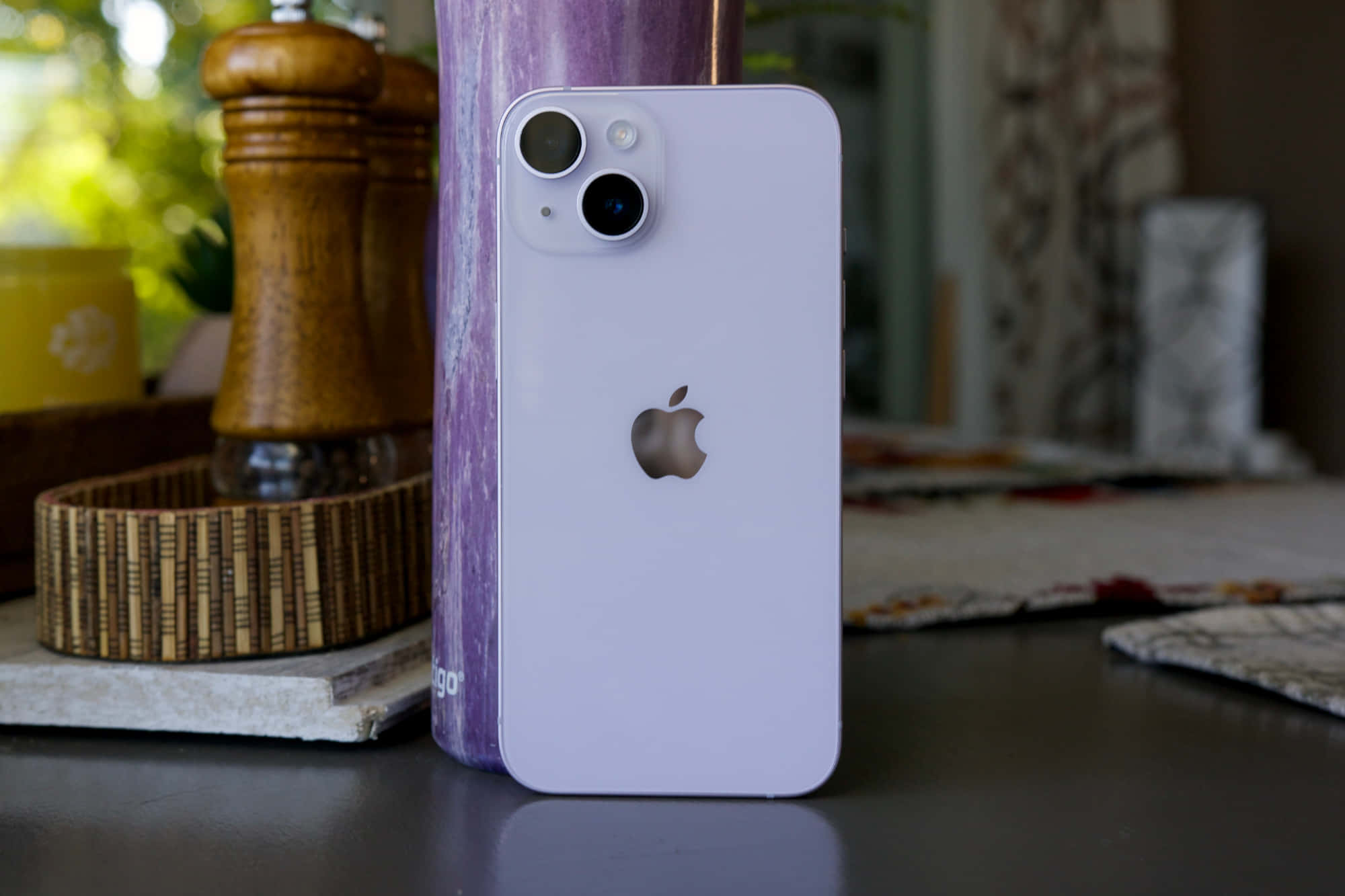 移動科技的下一個大突破：Apple Phone 壁紙 — 一部淺紫色iPhone 14直立放在一張深色桌子上，旁邊有一個木製胡椒磨和一個紫色花瓶，營造出舒適的廚房環境。