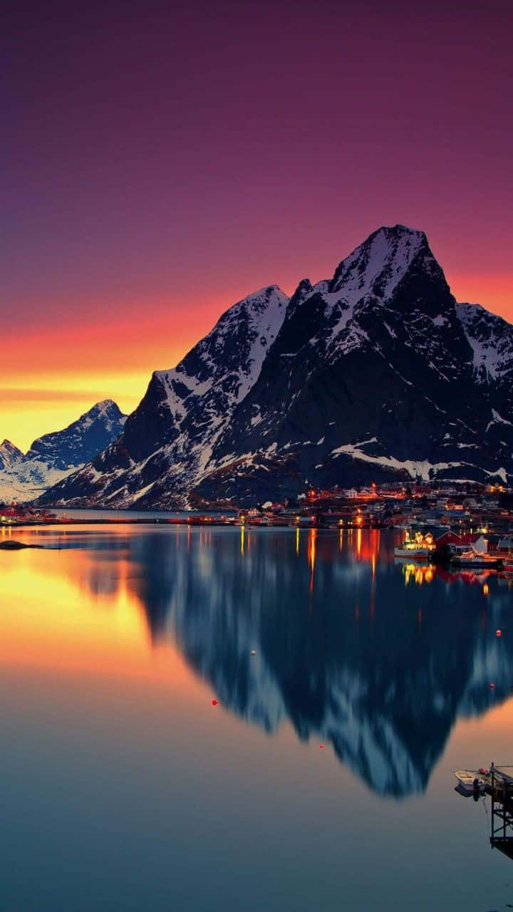 Norway 桌布 壁紙 — 積雪的山峰倒映在平靜的水面上，沿海村莊在充滿活力的紫色和橙色日落中閃耀。
