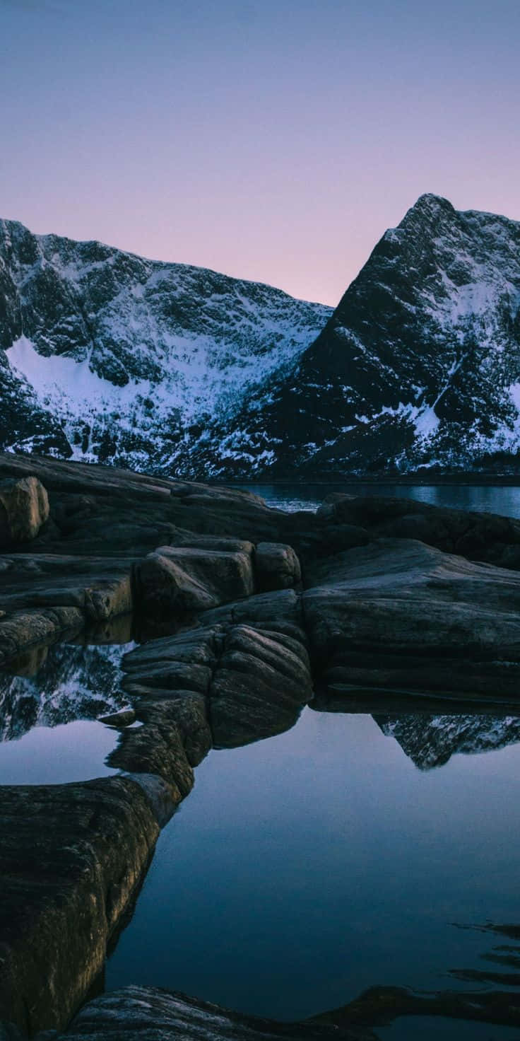 Norway 桌布 壁紙 — 積雪的山峰在暮色天空下，倒映在暗色岩石旁的平靜水面上。