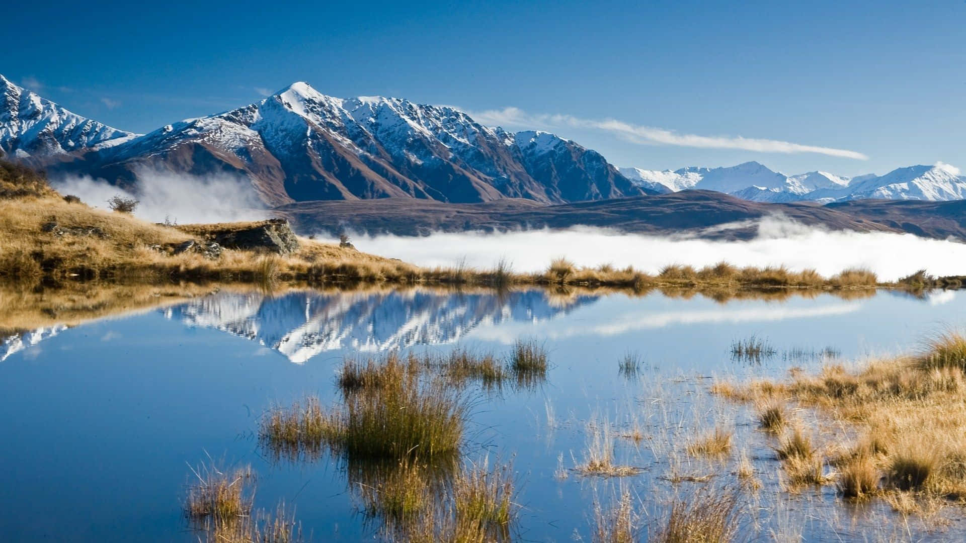 New Zealand 桌布 壁紙 — 積雪的山峰在霧氣瀰漫的山谷上方升起，完美地倒映在一個被金色草叢環繞的寧靜高山湖中。