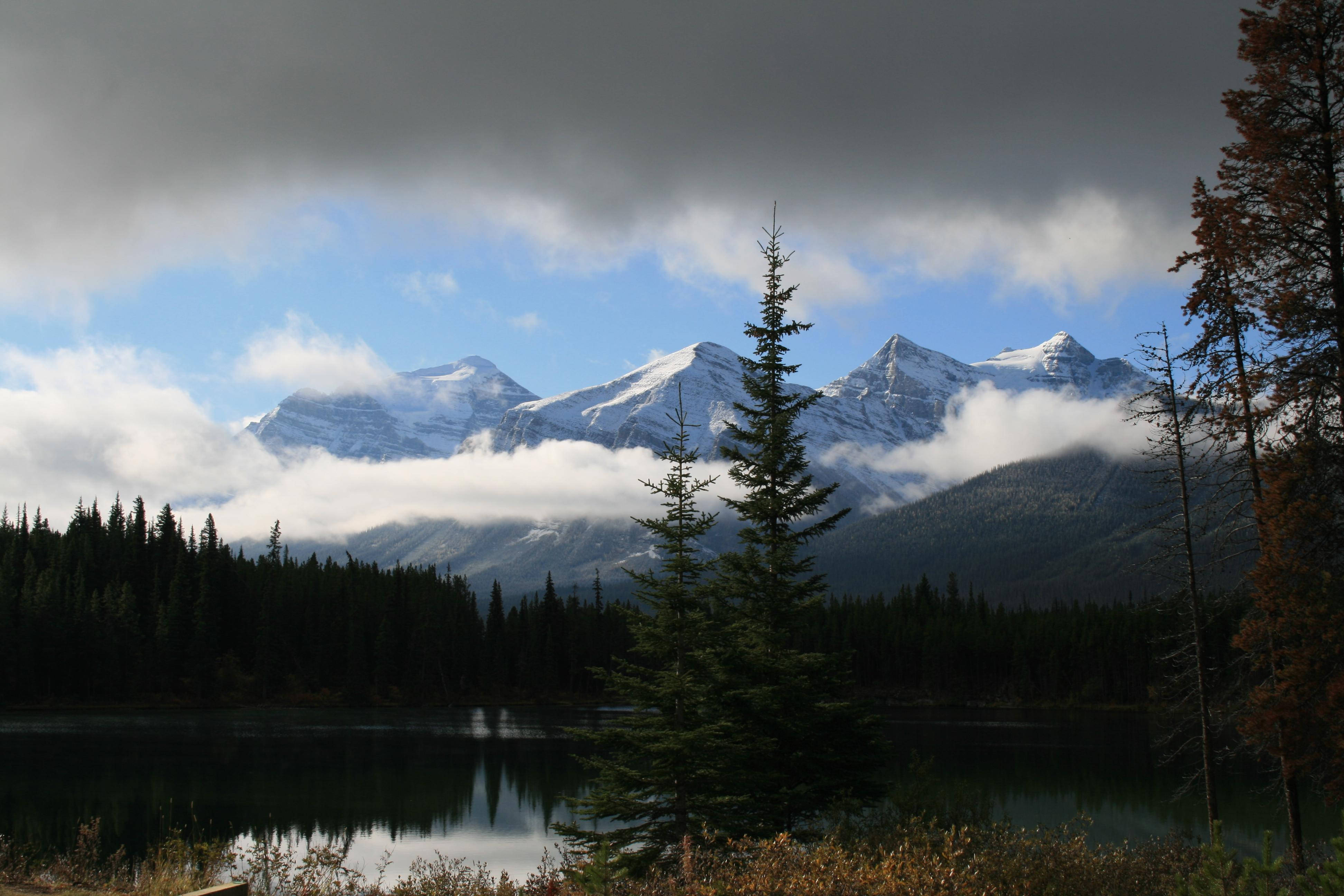 Mountain 桌布 壁紙 — 積雪覆蓋的山脈在平靜的湖泊後方升起，湖面倒映著雲朵和常綠樹，天空部分多雲。