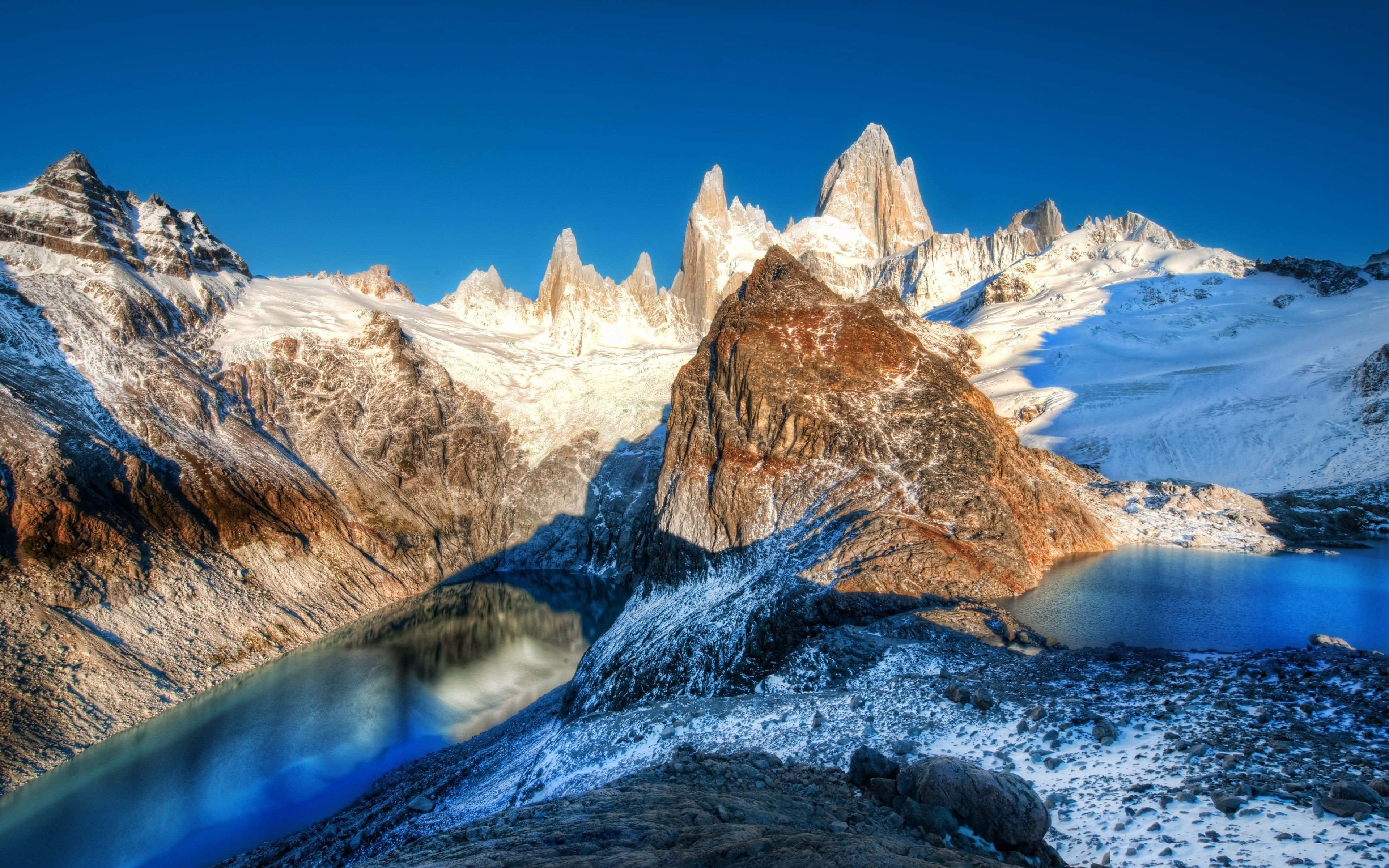 4K Hdr 桌布 壁紙 — 積雪覆蓋的鋸齒狀山峰刺破清澈的藍天，下方平靜的綠松石湖面倒映著山巒崎嶇的輪廓。