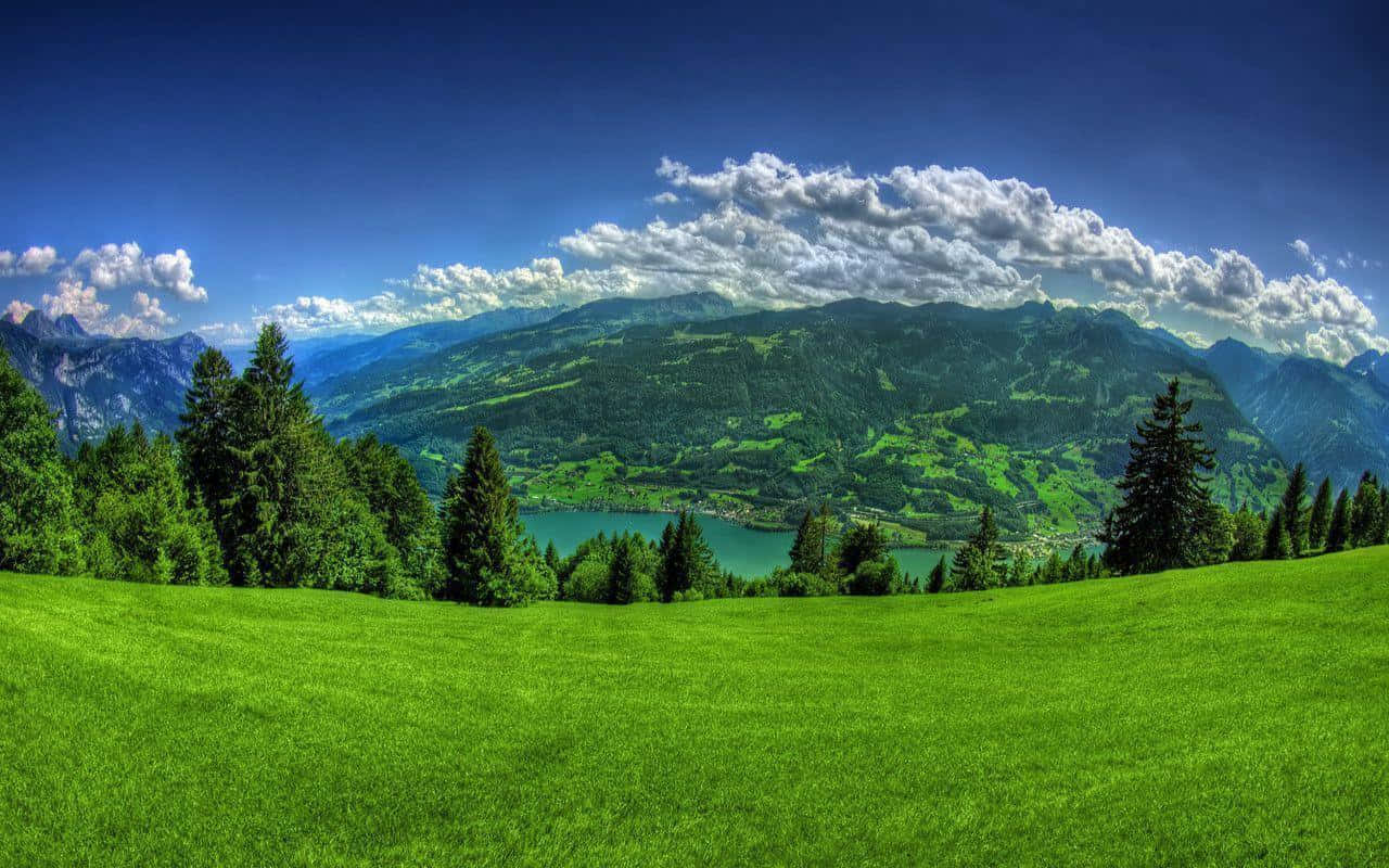 空中俯瞰的翠綠山景 壁紙 — 充滿活力的綠色草地向寧靜的湖泊傾斜，湖泊坐落在森林覆蓋的山脈之間，上方是明亮的藍天和蓬鬆的白雲。