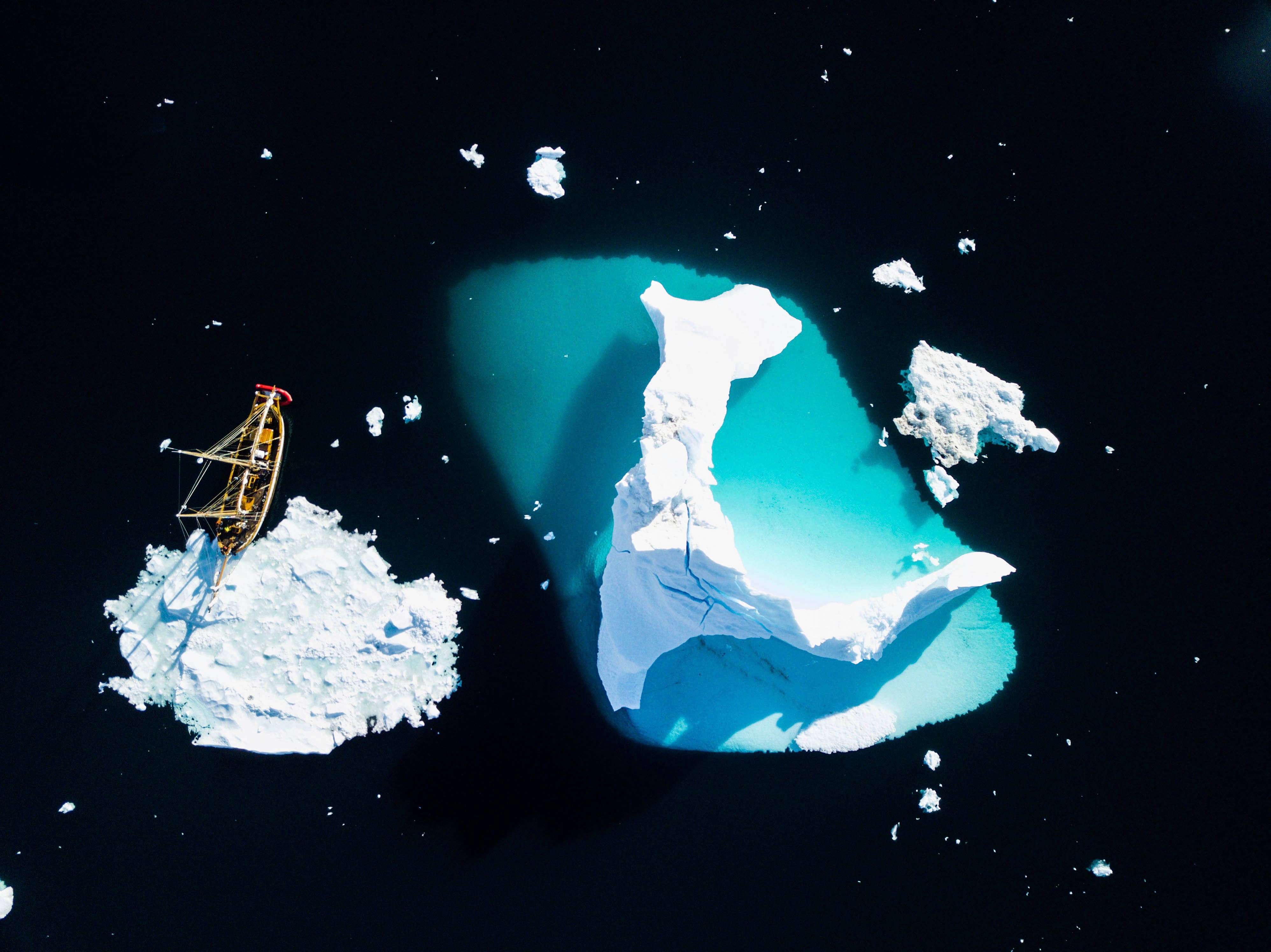 空中格陵蘭冰山 壁紙 — 從高空俯瞰，一艘小船停泊在白雪覆蓋的冰山上，旁邊是巨大的藍綠色浮冰，漂浮在黑暗的海水中。