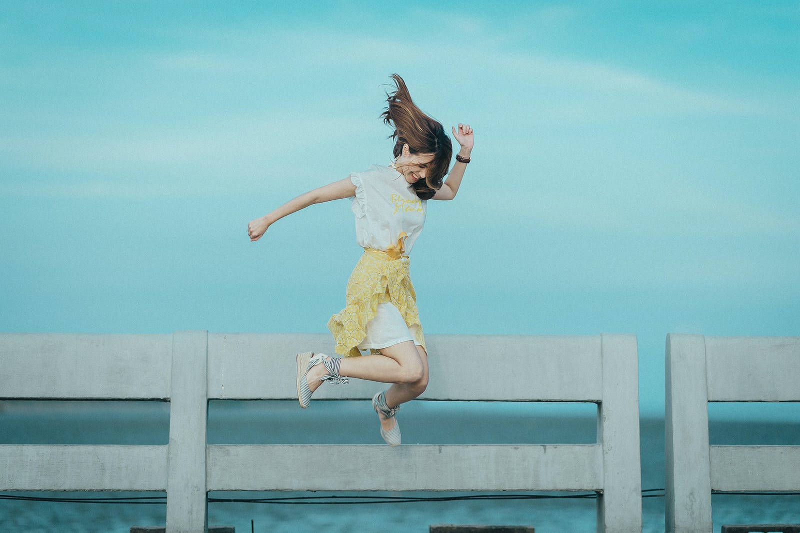 空中舞蹈的自由時刻 壁紙 — 一個快樂的女孩在寧靜的綠松色天空和海洋背景下，飛躍過混凝土欄杆。