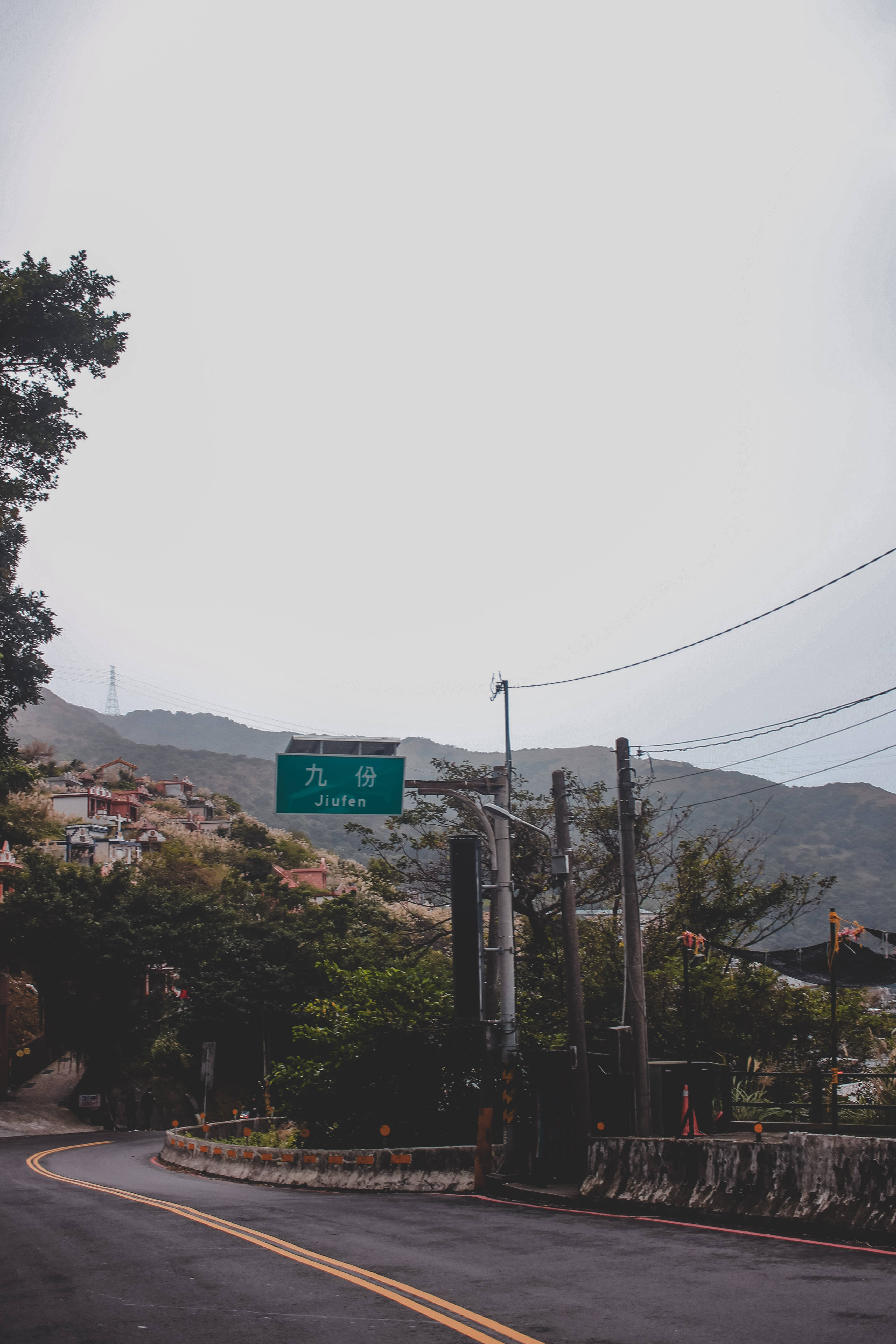 空曠街景美學鎖屏 壁紙 — 一條蜿蜒的道路穿過霧氣瀰漫的山村，瀝青路面上方懸掛著一個綠色的 Jiafen 標誌。