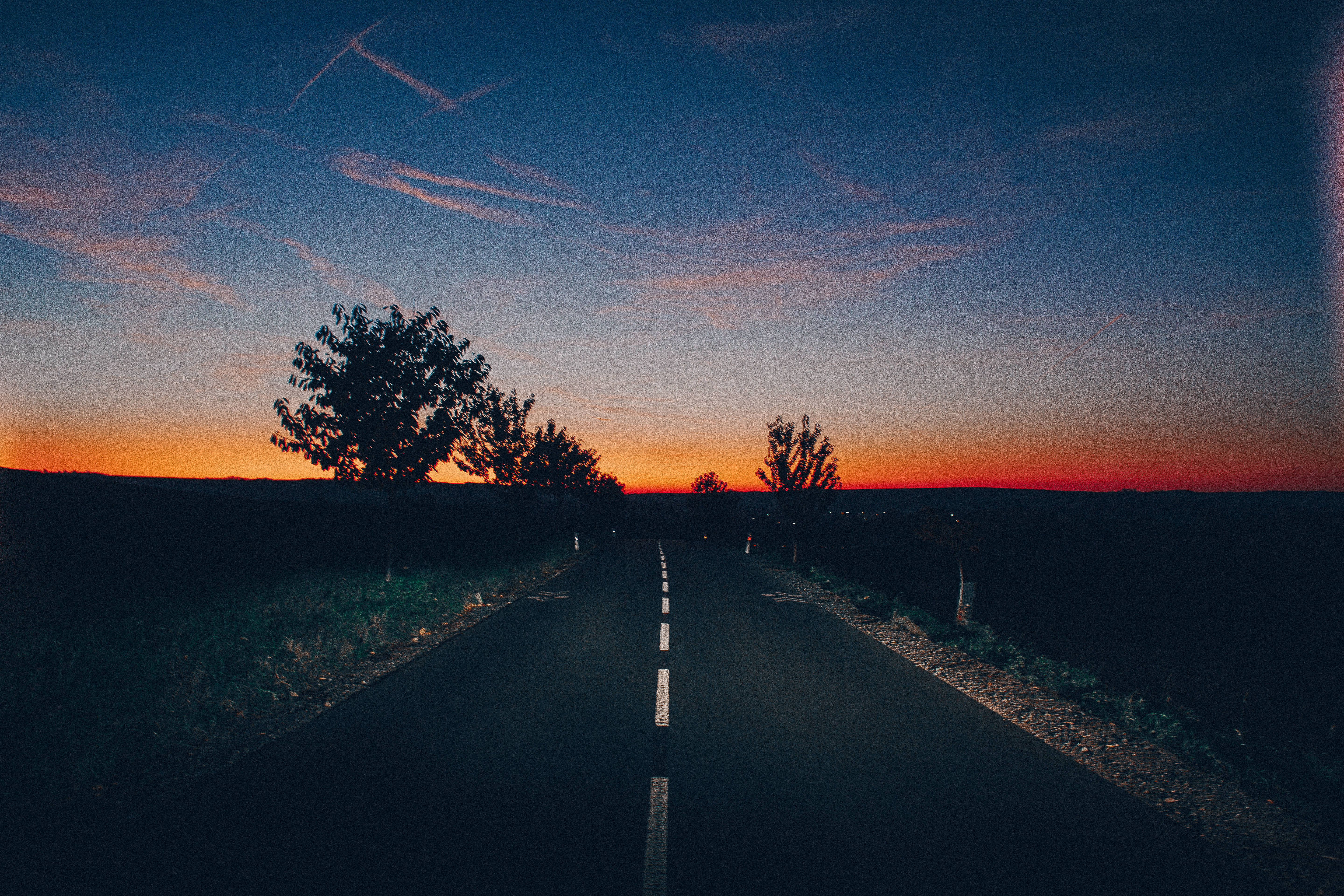 空曠道路與樹木上方的夜空美學壁紙 壁紙 — 一條孤獨的道路在暮色天空下延伸至地平線，天空染上熾熱的橙色和深藍色調。