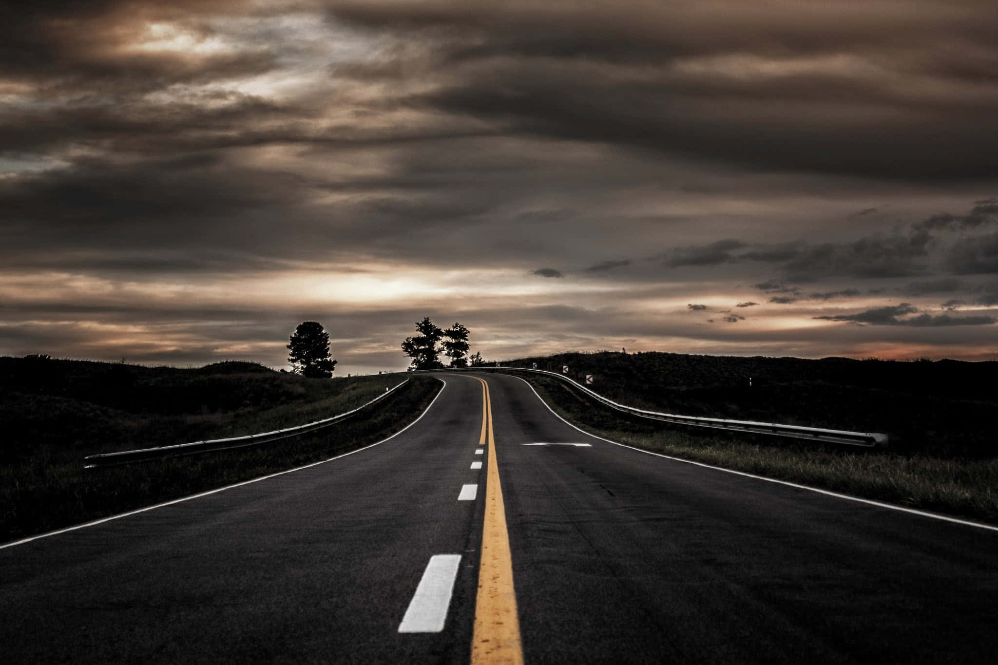 空曠道路與深色天空 壁紙 — 一條蜿蜒的瀝青道路在黃昏時分戲劇性的多雲天空下延伸至遠方，地平線上有樹木的剪影。