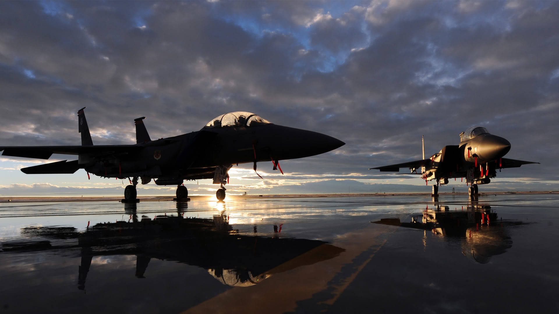 空軍 F-15 壁紙 — 兩架戰鬥機在濕滑的停機坪上形成剪影，夕陽在它們身後落下，投射出戲劇性的倒影。