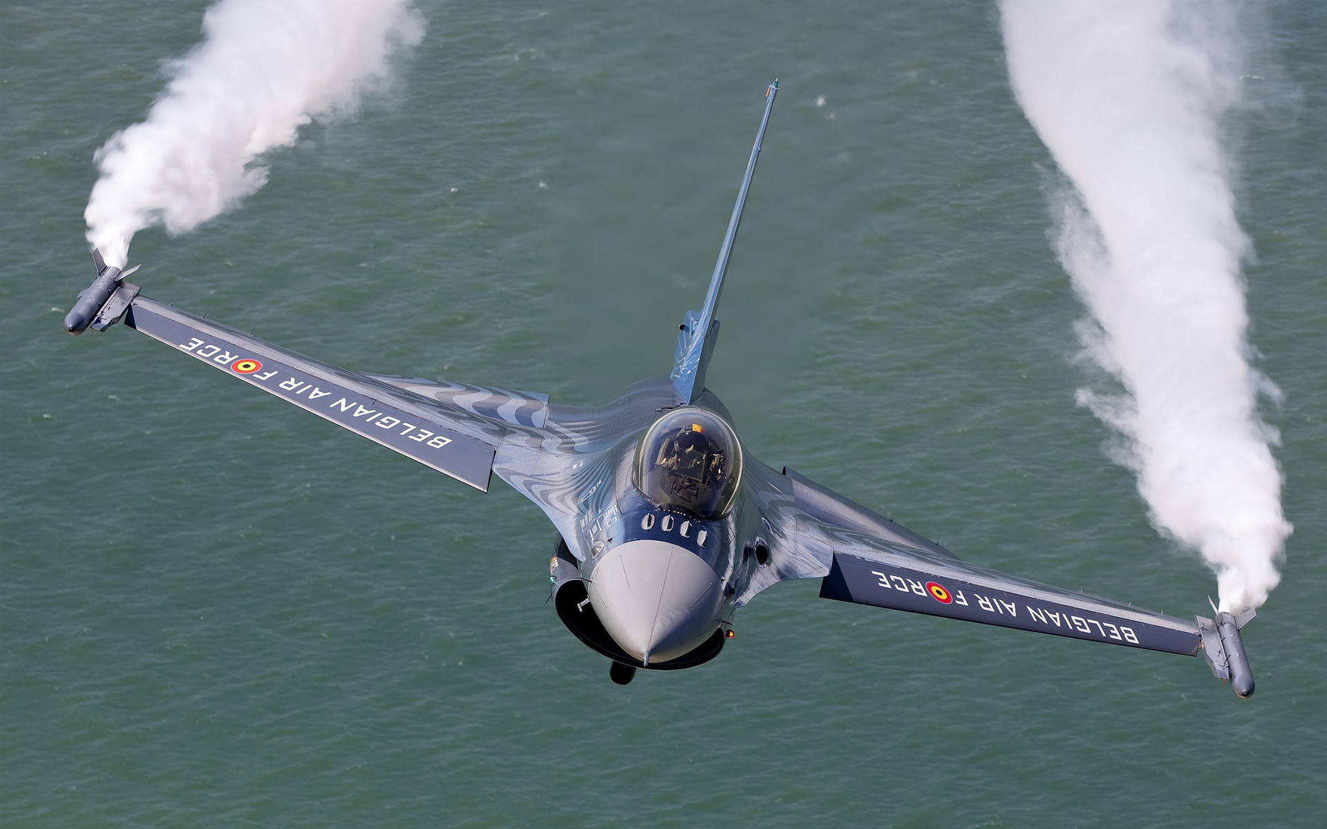 空軍戰機尾煙 壁紙 — 一架比利時空軍噴射機低飛掠過海洋水面，機翼末端拖出濃厚白色煙霧。