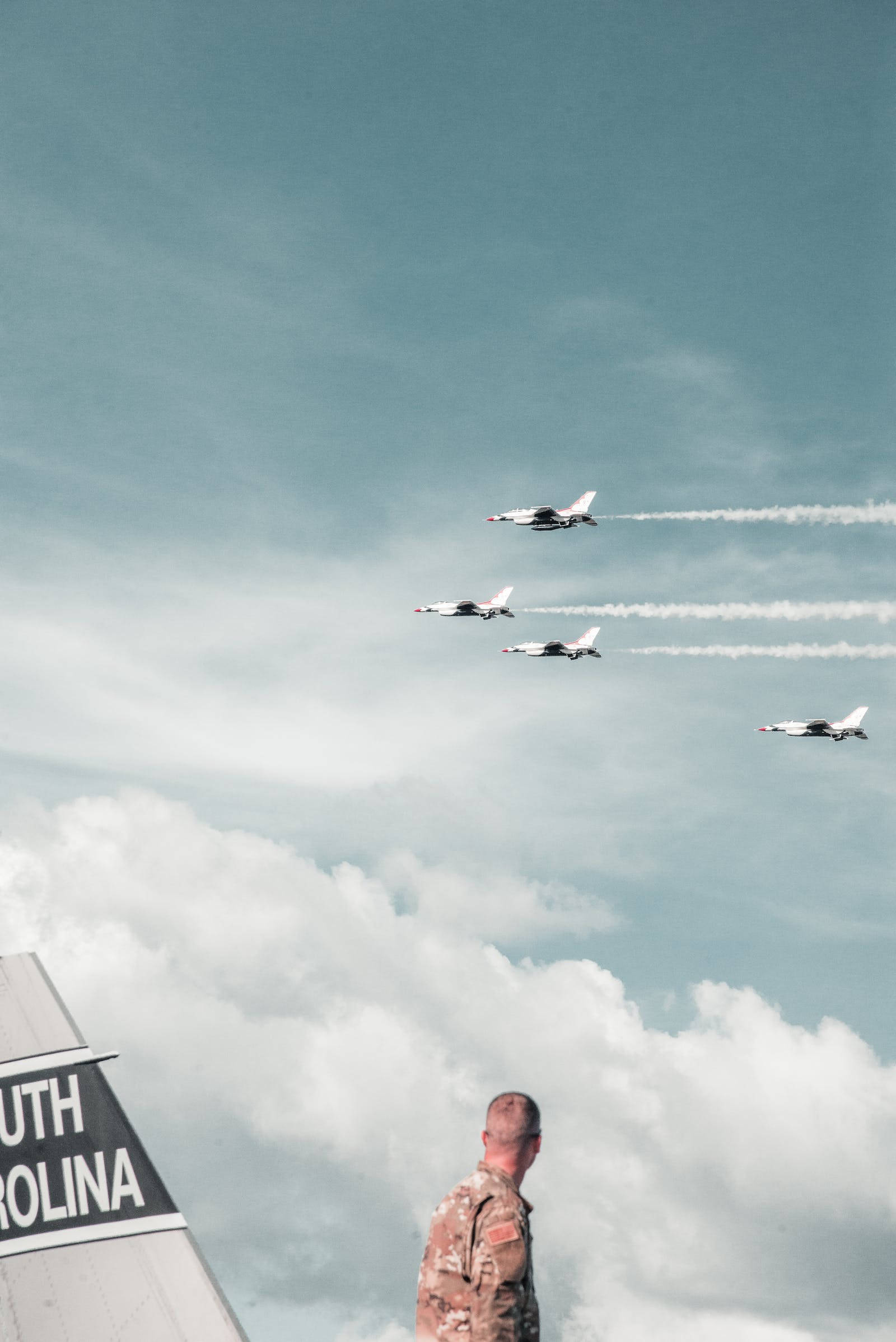 空軍戰鬥機南卡羅來納州 壁紙 — 一名身穿迷彩服的士兵站立觀看四架戰鬥機在頭頂編隊飛行，在陰雲密佈的天空中留下白色煙跡。