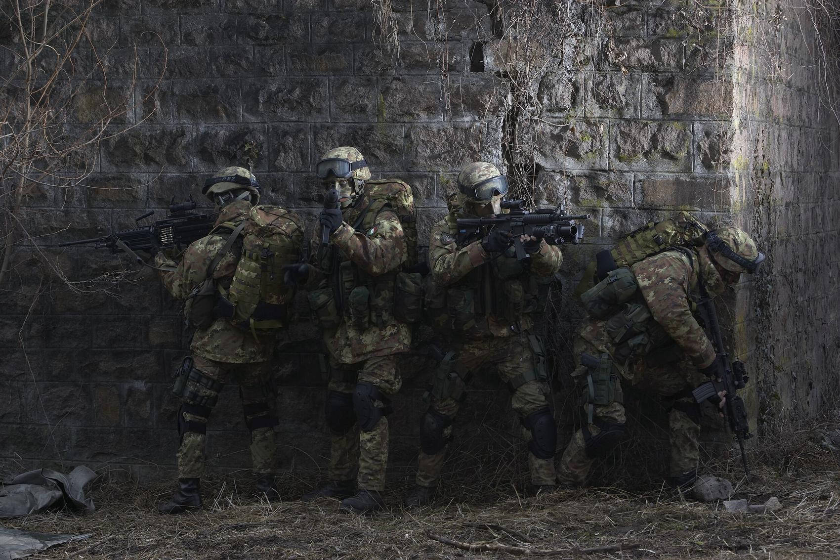 四名身穿迷彩裝備的武裝士兵蹲在石牆後面，處於戰鬥姿態。