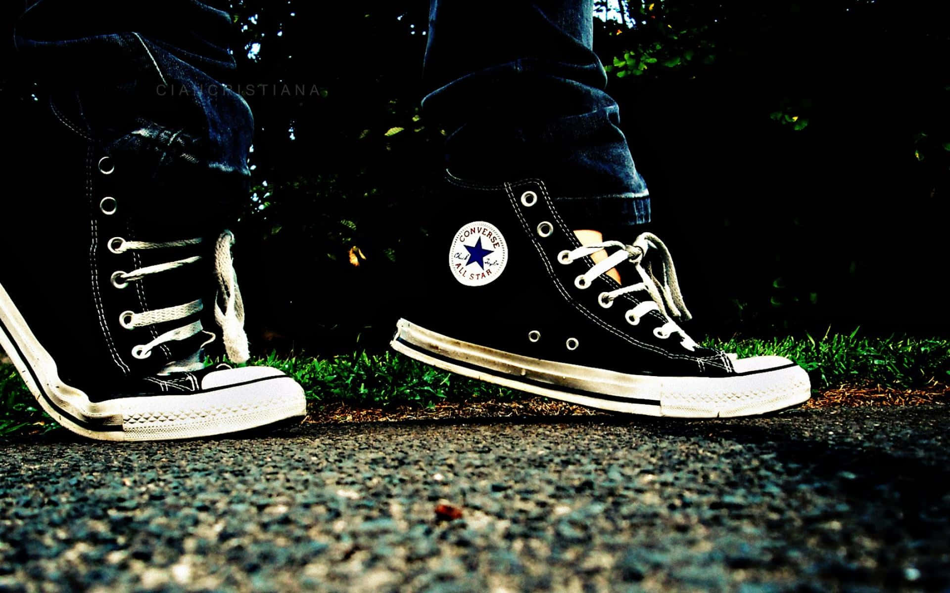 穿Converse，自信每一步 壁紙 — 磨損的黑色Converse運動鞋特寫，帶白色鞋帶和鞋底，站在暗色樹葉下的草地附近的礫石上。