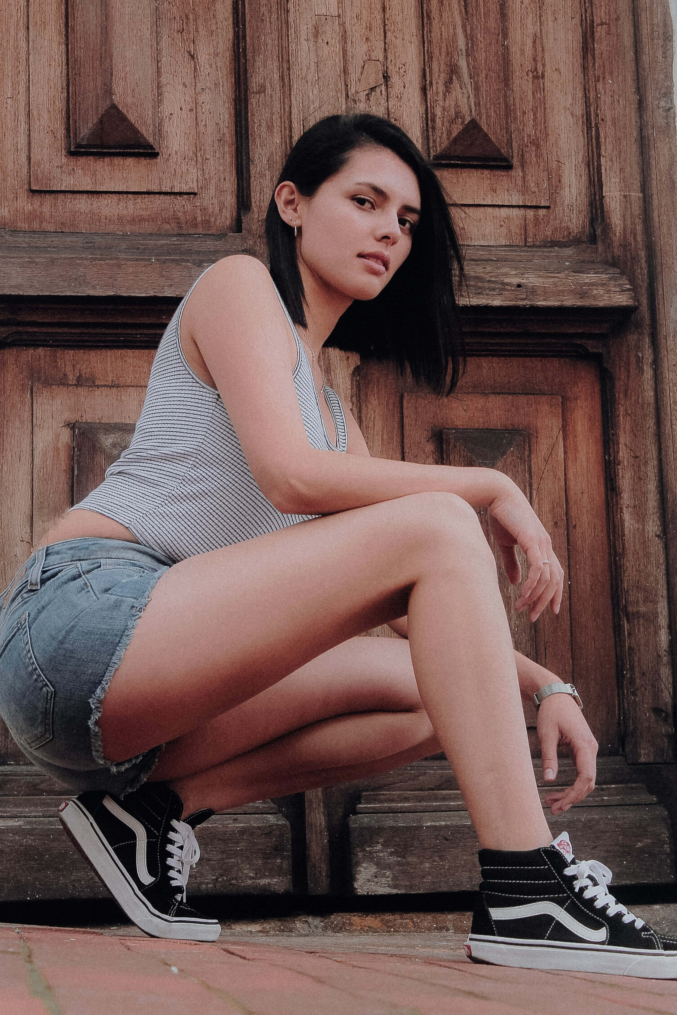 穿Vans高幫鞋展現修長美腿的女性 壁紙 — 一位身穿牛仔短褲和黑色運動鞋的年輕女子自信地蹲在風化的木門前。