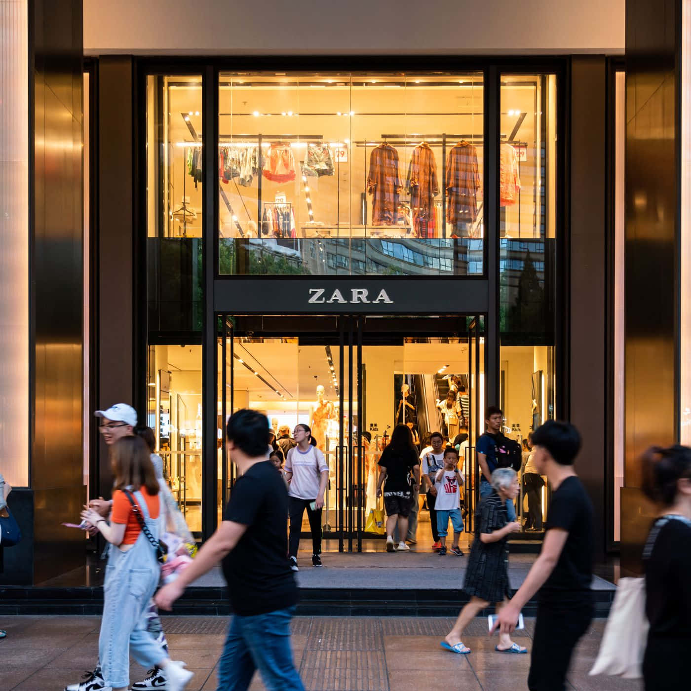 穿Zara，好看又舒適 壁紙 — 行人經過一間燈火通明的Zara商店，透過大型玻璃窗可以看到服裝展示。