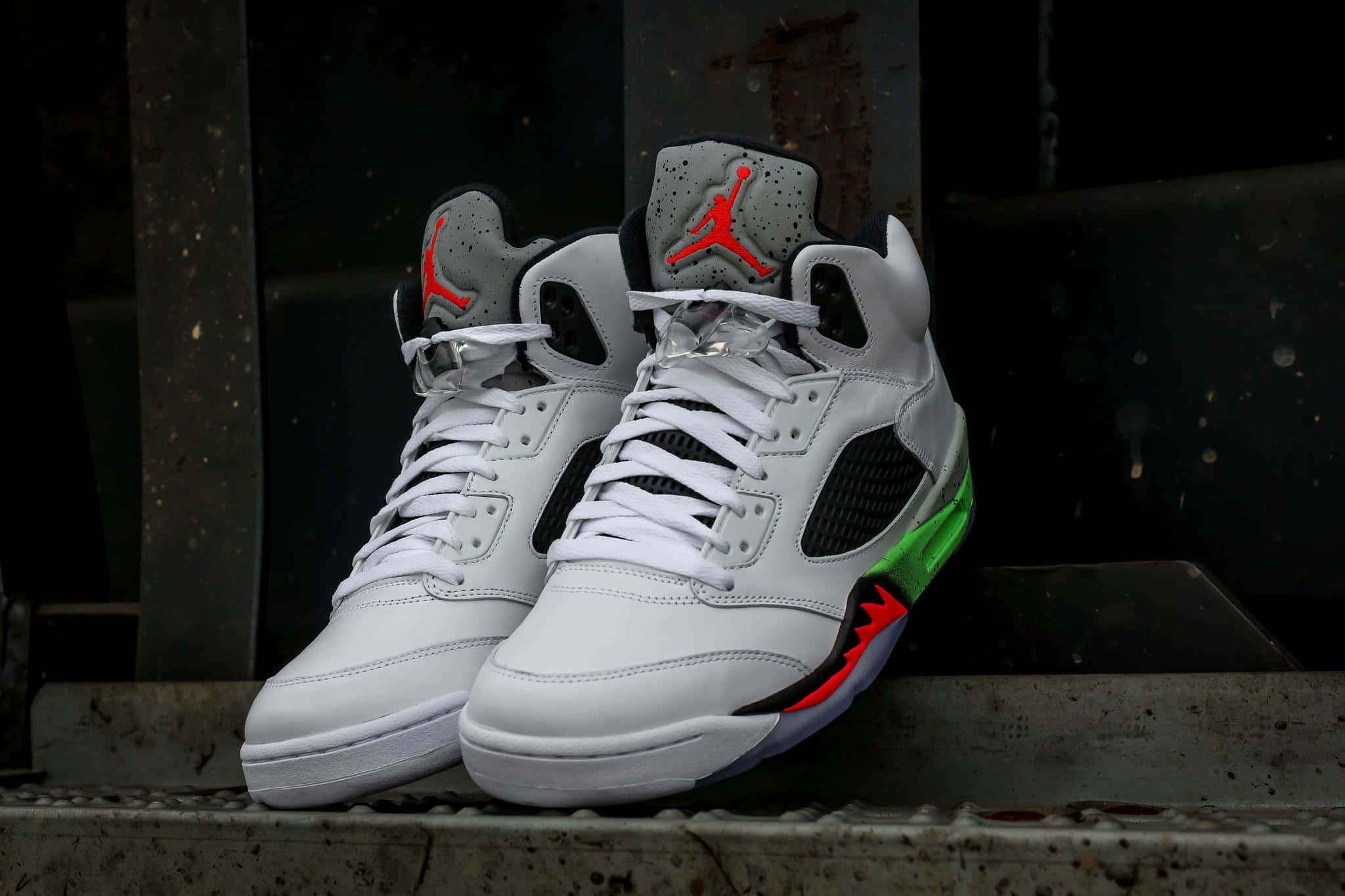 穿上Air Jordan，提升你的時尚品味 壁紙 — 一對白色Jordan 5運動鞋，帶有紅色和綠色點綴，放在風化的混凝土邊緣上，背景是黑暗的工業環境。