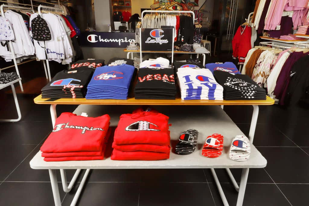 穿上冠軍的冠軍造型 壁紙 — 整齊堆疊的Champion品牌服裝和鞋類展示在零售店內的白色桌子上，背景有衣架。