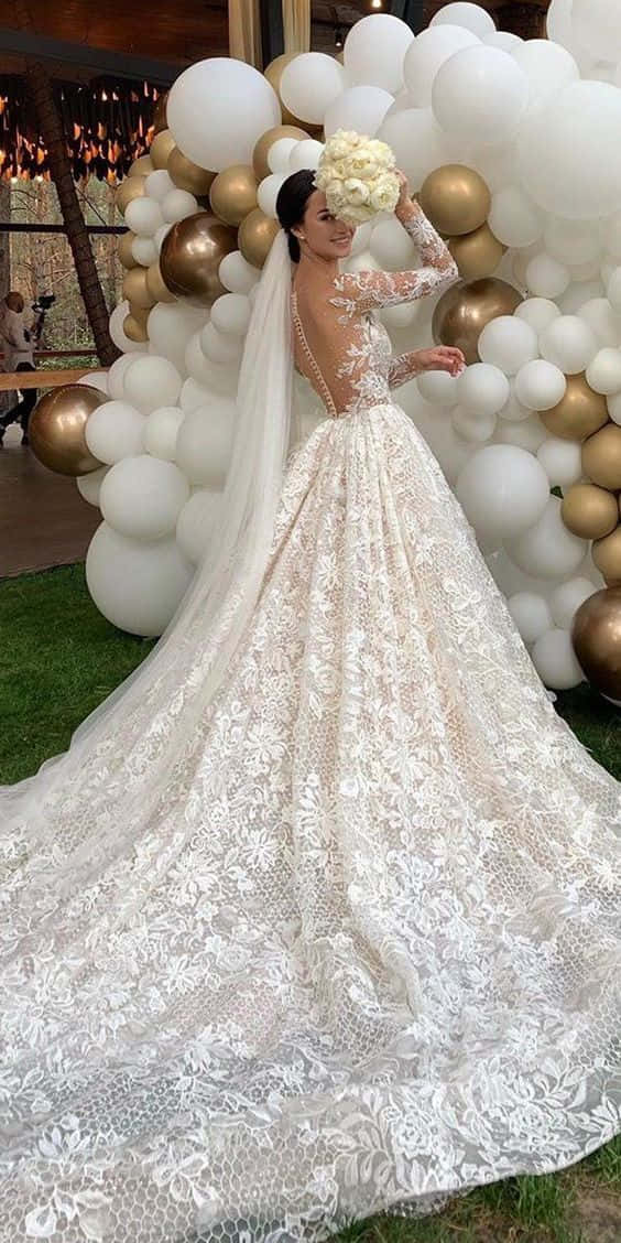 穿上這款優雅婚紗，顯得無與倫比。 壁紙 — 一位身穿精緻白色蕾絲婚紗、長長拖尾的新娘，站在白色和金色氣球牆前，手持花束。