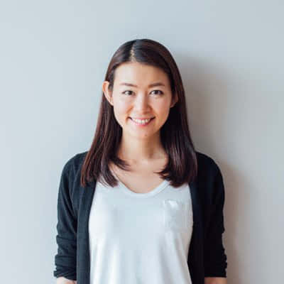一位棕色頭髮的女性，身穿白色襯衫和黑色開襟毛衣，在灰色牆前微笑。
