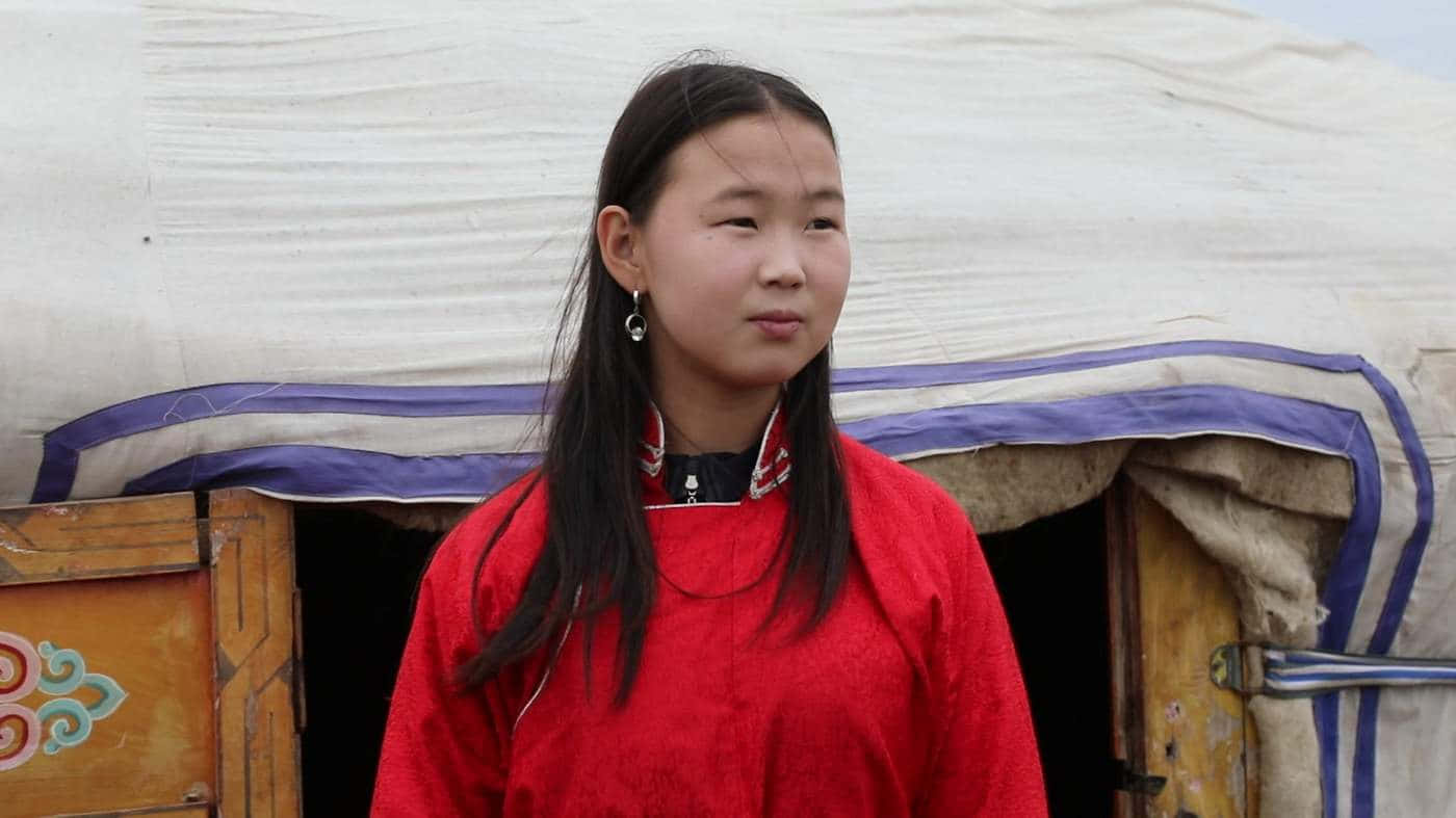 穿傳統蒙古服裝的亞洲美女 壁紙 — 一位年輕女孩穿著鮮豔的紅色傳統服裝，站在一扇有木門和裝飾圖案的風化蒙古包前。