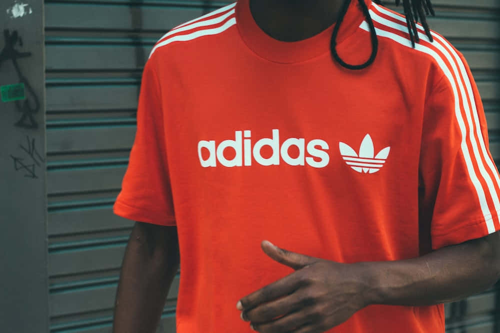 穿得好看，穿得舒服 — #adidas 壁紙 — 一個人穿著一件大膽的橙色adidas T恤，帶有白色條紋和標誌，背景是塗鴉噴漆的金屬百葉窗。