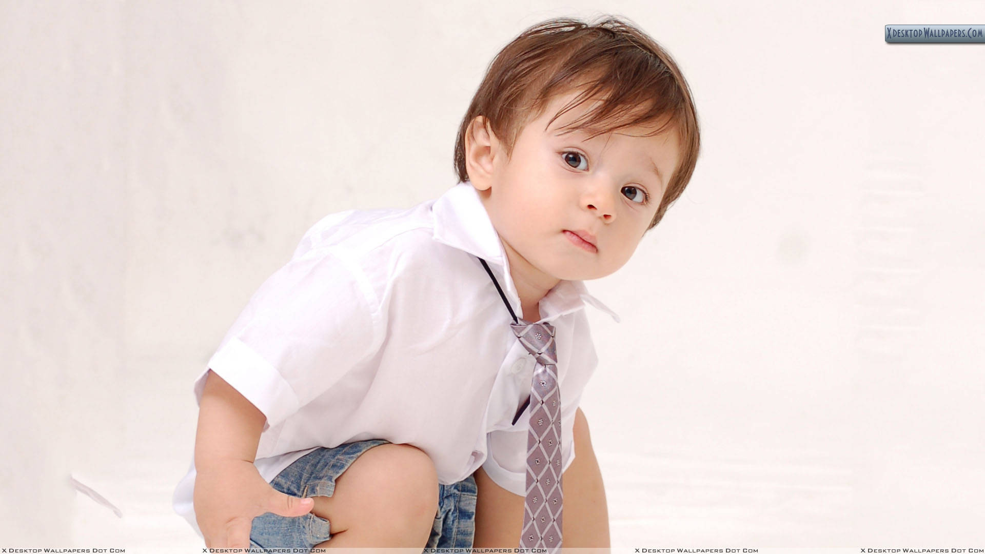 穿時尚裝束的男孩 壁紙 — 一個好奇嘅幼兒，著住白色恤衫同圖案領呔，坐喺純白背景前，表情認真。