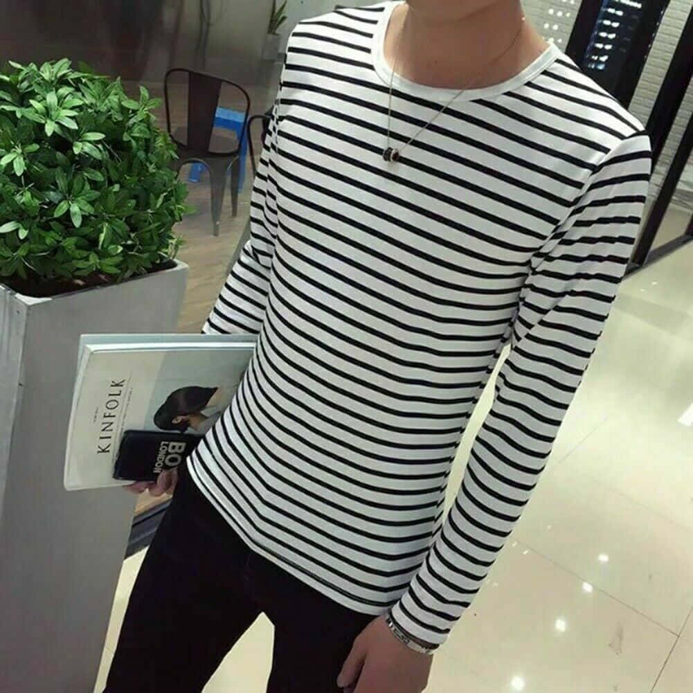 穿條紋上衣的男性模特 壁紙 — 一位年輕男子穿著黑白條紋長袖襯衫，在現代室內環境中拿著雜誌靠近盆栽植物。