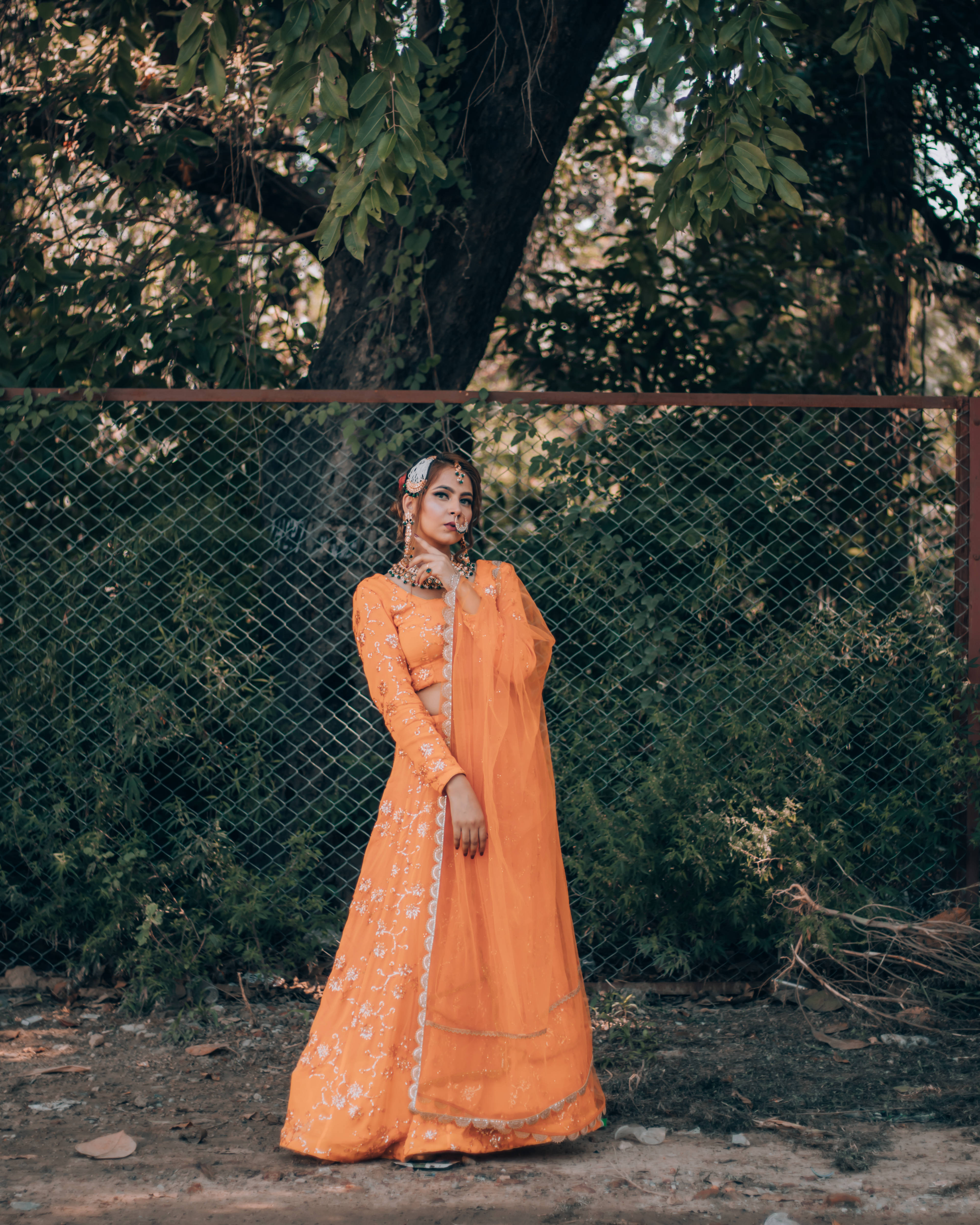 穿橙色服裝的印度風女性 壁紙 — 一位身穿鮮豔橙色刺繡 lehenga 的女性，在樹葉茂密的樹木過濾下的斑駁陽光中，優雅地站在鐵絲網圍欄旁。