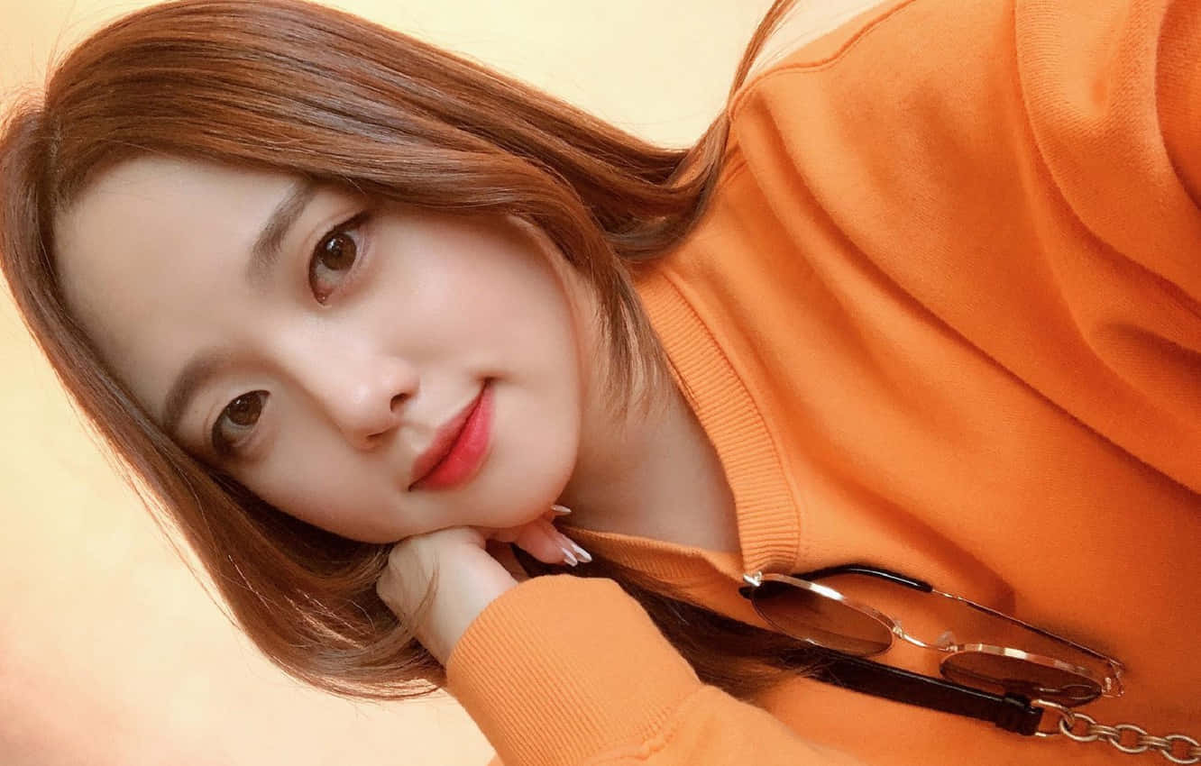穿橙色長袖上衣的可愛韓國女孩 壁紙 — 一位棕色頭髮的年輕女子，身穿橙色毛衣，太陽眼鏡掛在肩上，手托著下巴。