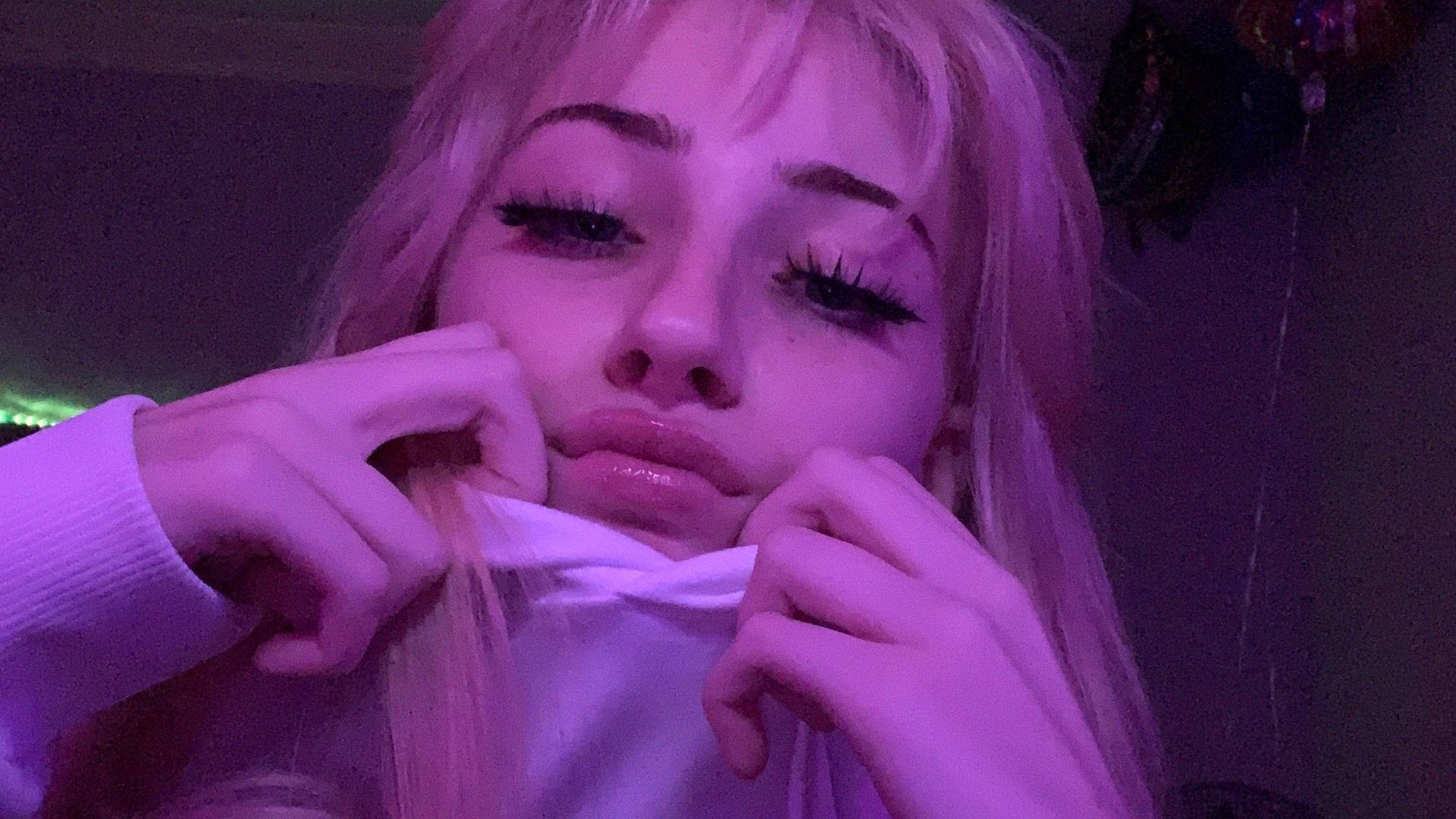 穿毛衣的Baddie風格女孩 壁紙 — 一位擁有金色長髮和戲劇化妝容的人，在紫色燈光下拉扯白色襯衫的領口。