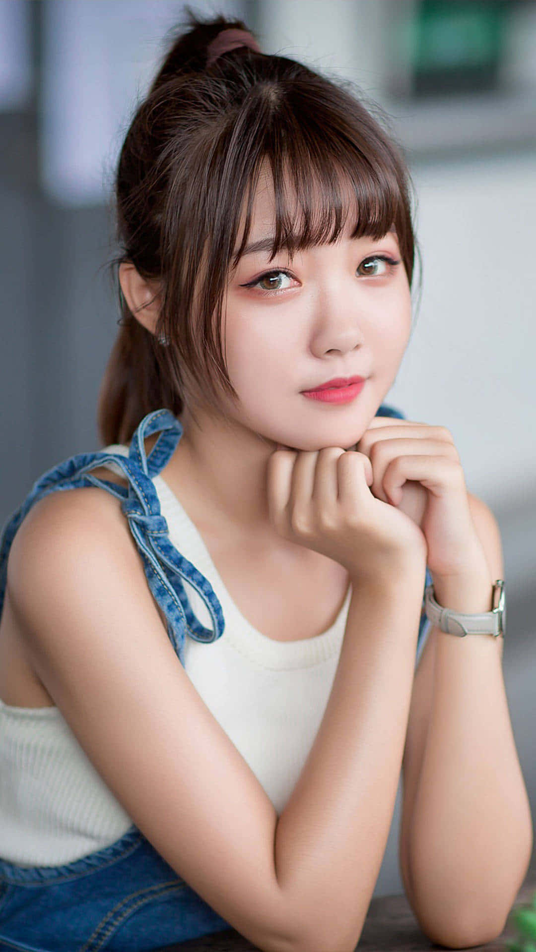 穿牛仔背心的可愛韓國女孩 壁紙 — 一位年輕女性紮著棕色馬尾辮，身穿牛仔背帶褲和白色上衣，雙手托腮。