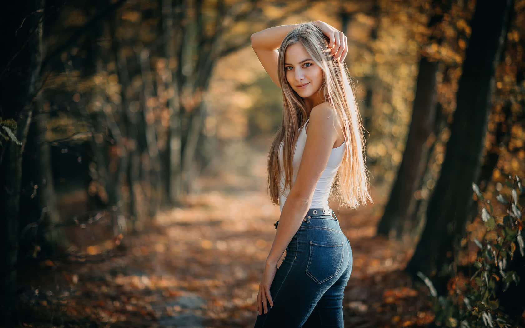 穿牛仔褲的森林女孩 壁紙 — 一位長金髮的女性在陽光明媚的秋林小徑上回頭微笑。