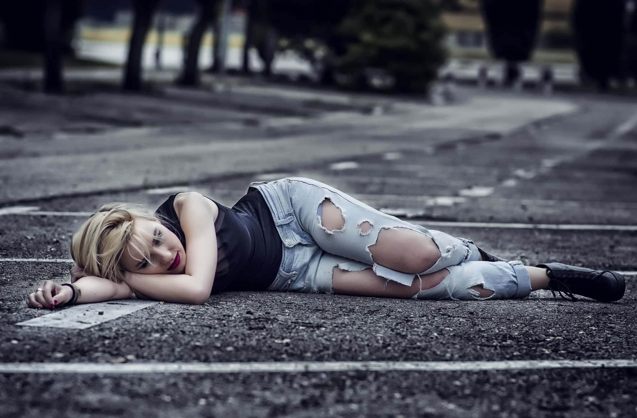 穿牛仔褲躺路邊的女孩圖 壁紙 — 一位金髮年輕女子側臥在鋪砌的道路上，穿著破洞牛仔褲和黑色上衣。