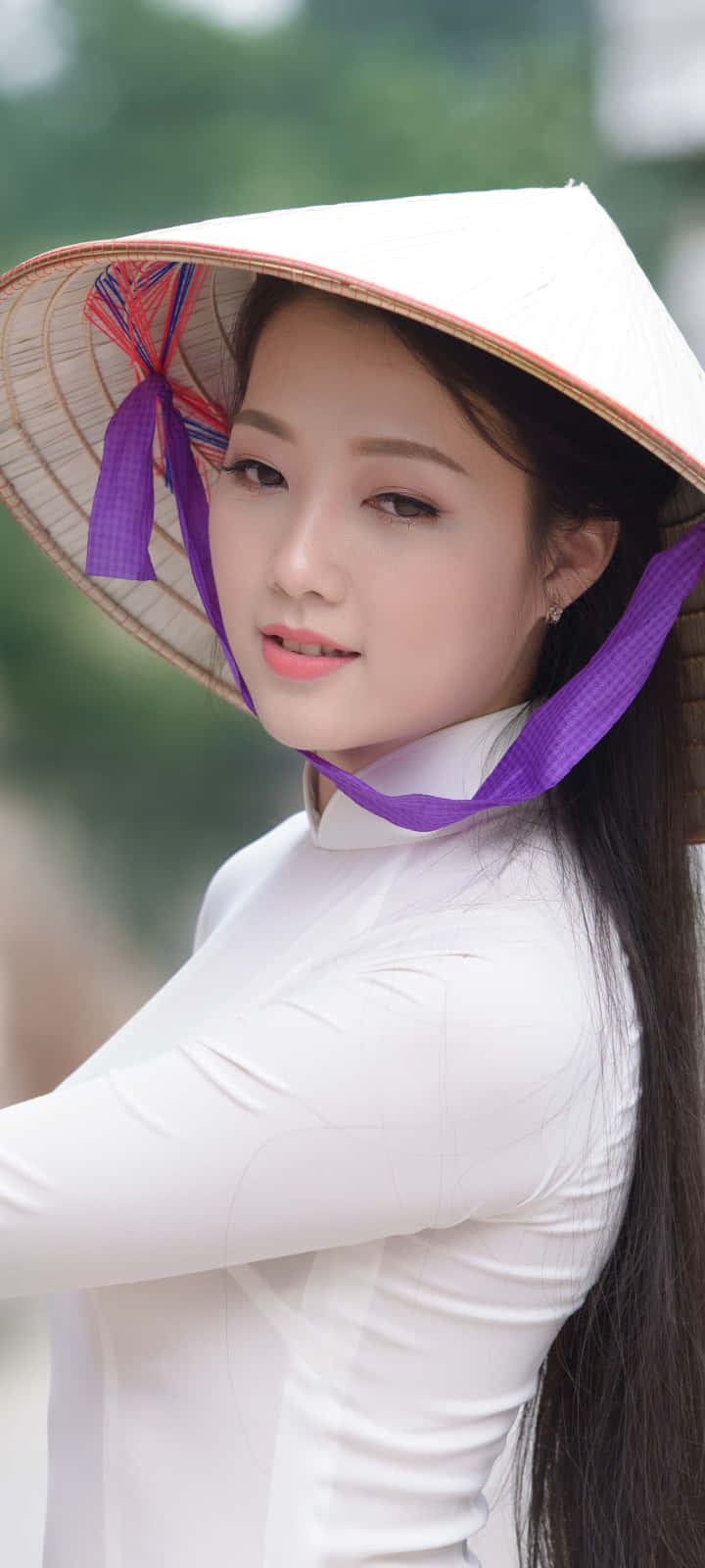 穿白色禮服的亞洲女性 壁紙 — 一位身穿白色奧黛和傳統錐形帽（帶紫色絲帶）的年輕女性，在模糊的綠色背景下溫柔地凝視。