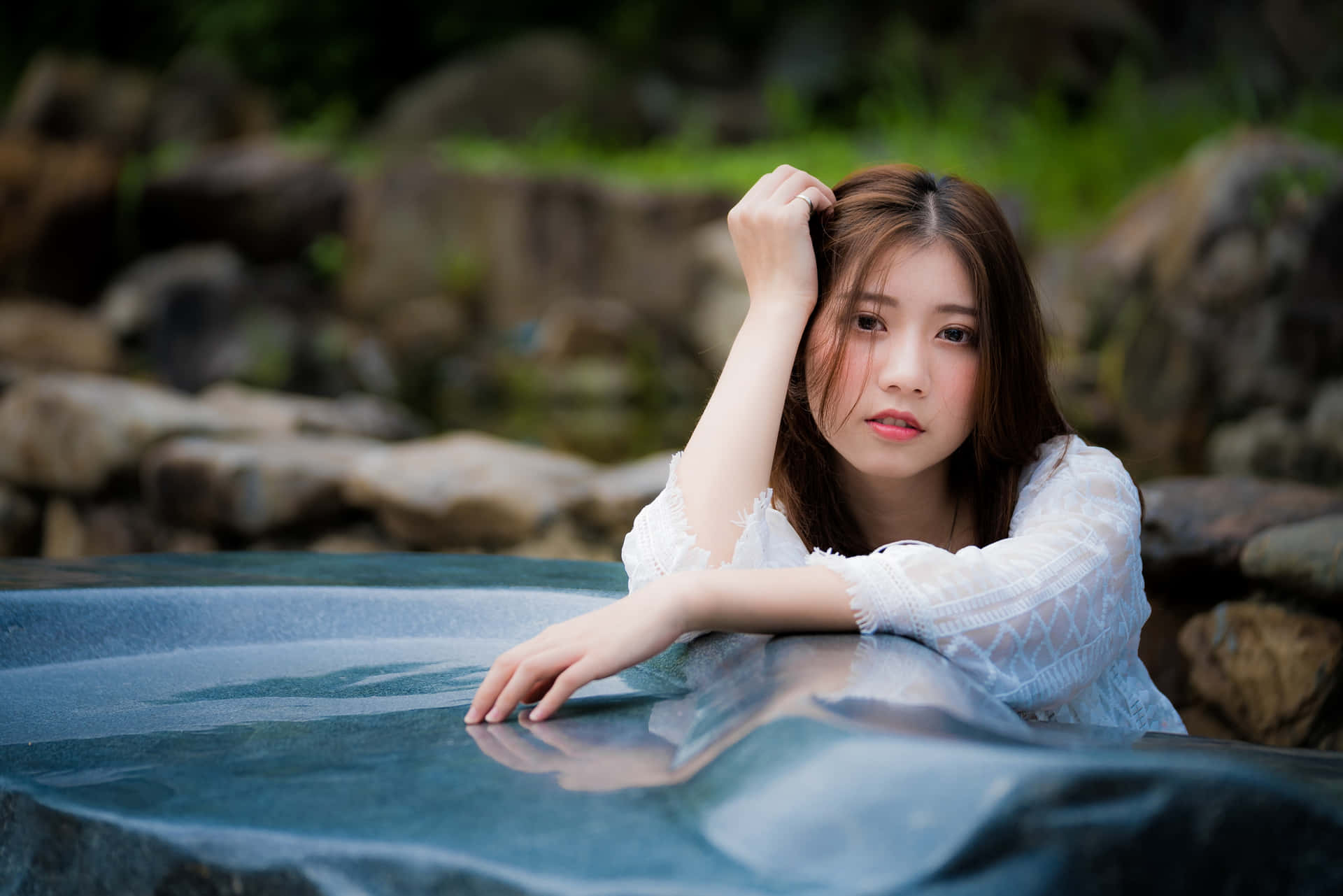 年輕女子身穿白色蕾絲上衣，在自然環境中將手臂擱在反射水池上