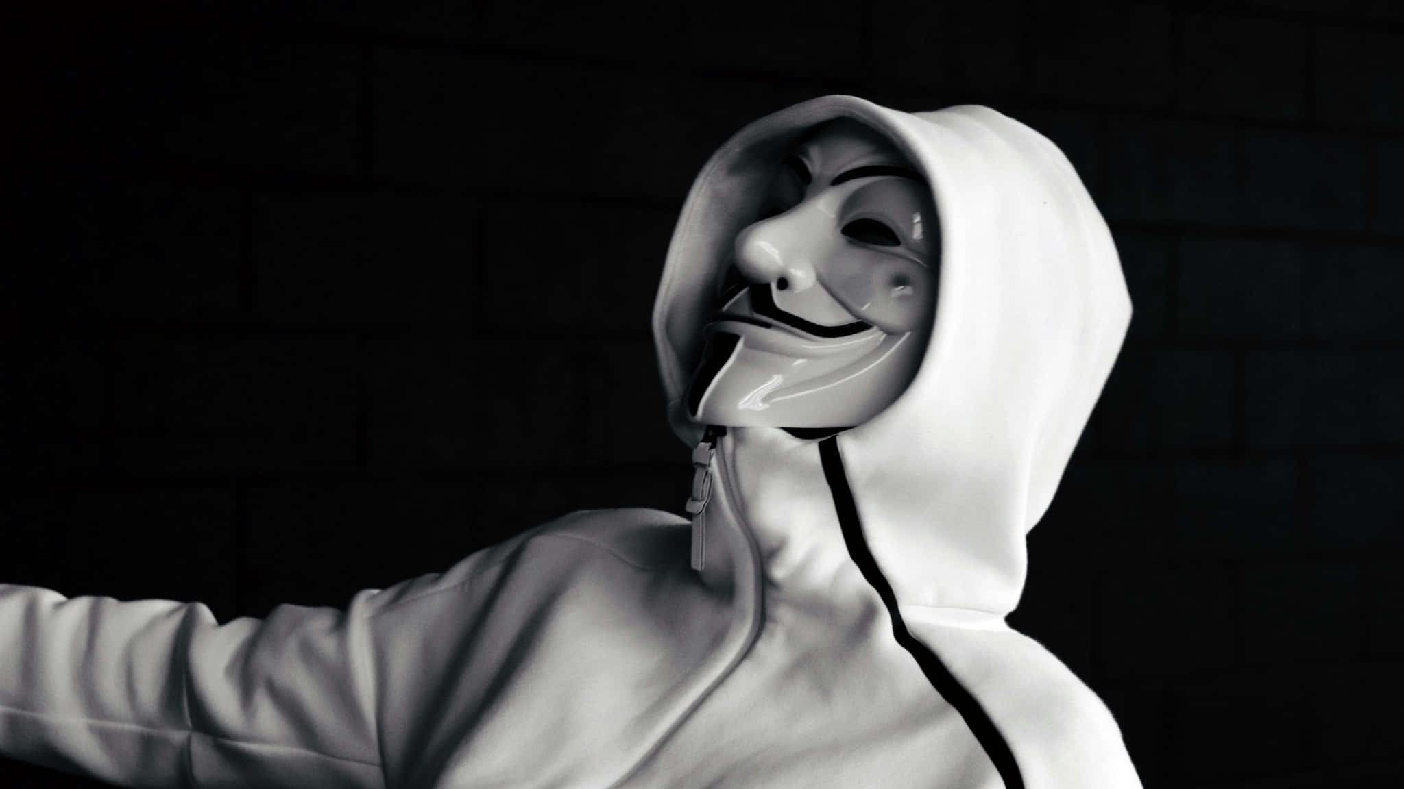 穿白色運動服的 Vendetta 面具男子漂浮空中 壁紙 — 一名穿著白色連帽衫的人，在戲劇性燈光下，戴著蓋伊·福克斯面具，背景是深色磚牆。