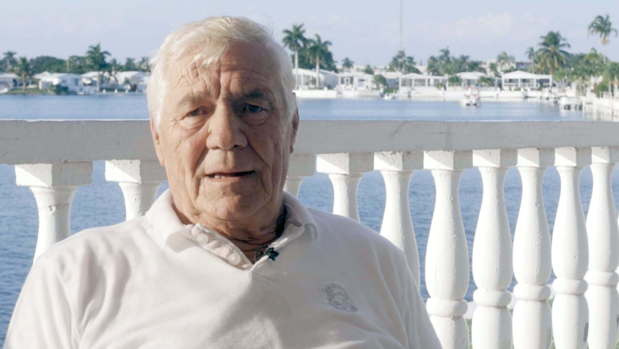 穿白襯衫的傳奇摔角手 Pat Patterson 壁紙 — 一位白髮老人坐在白色陽台上，俯瞰著寧靜的海濱，遠處有棕櫚樹和房屋。