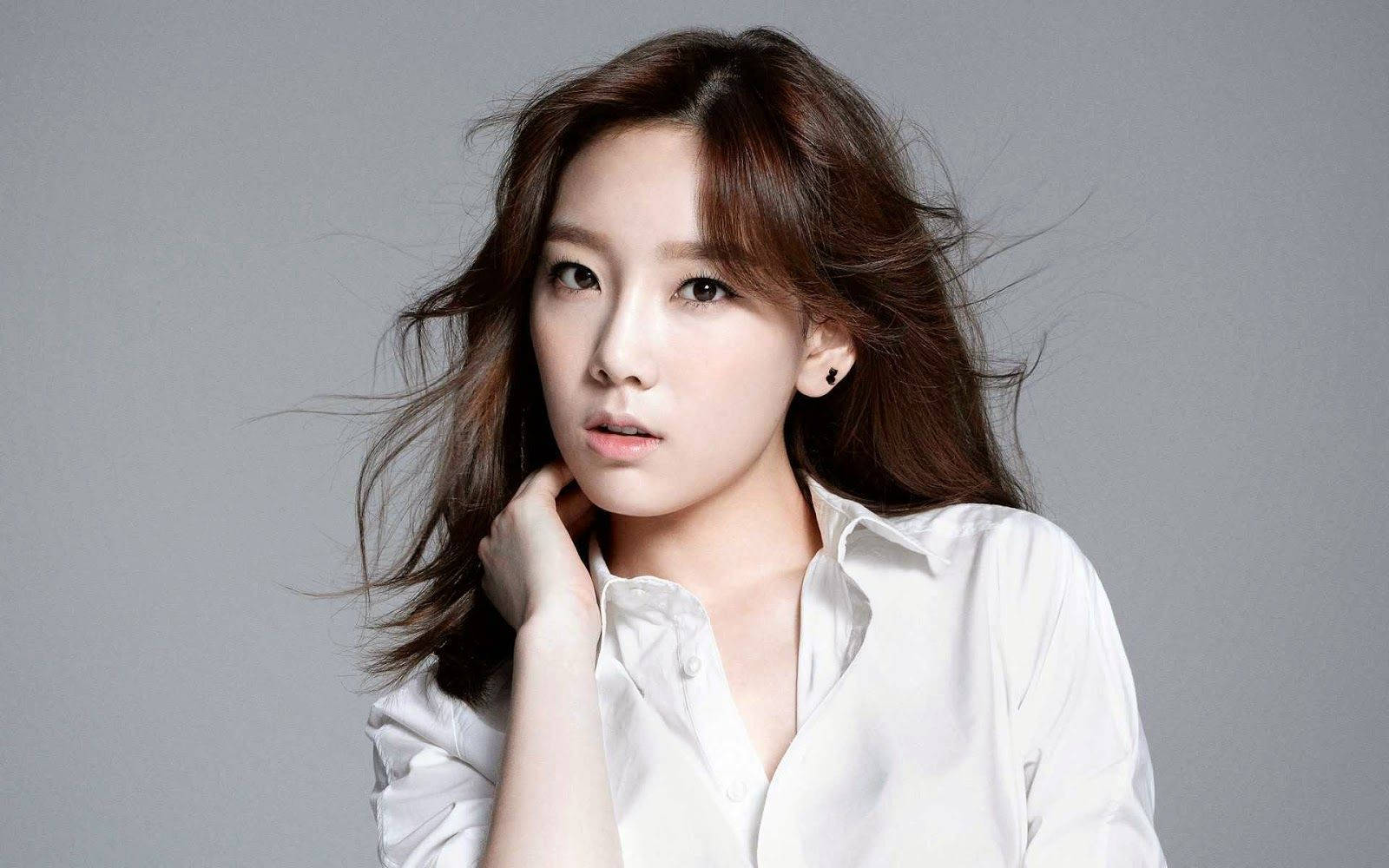 穿白襯衫的酷女孩Taeyeon 壁紙 — 一位年輕女性，棕髮隨風飄揚，身穿白色襯衫，在純灰色工作室背景前若有所思地凝視。
