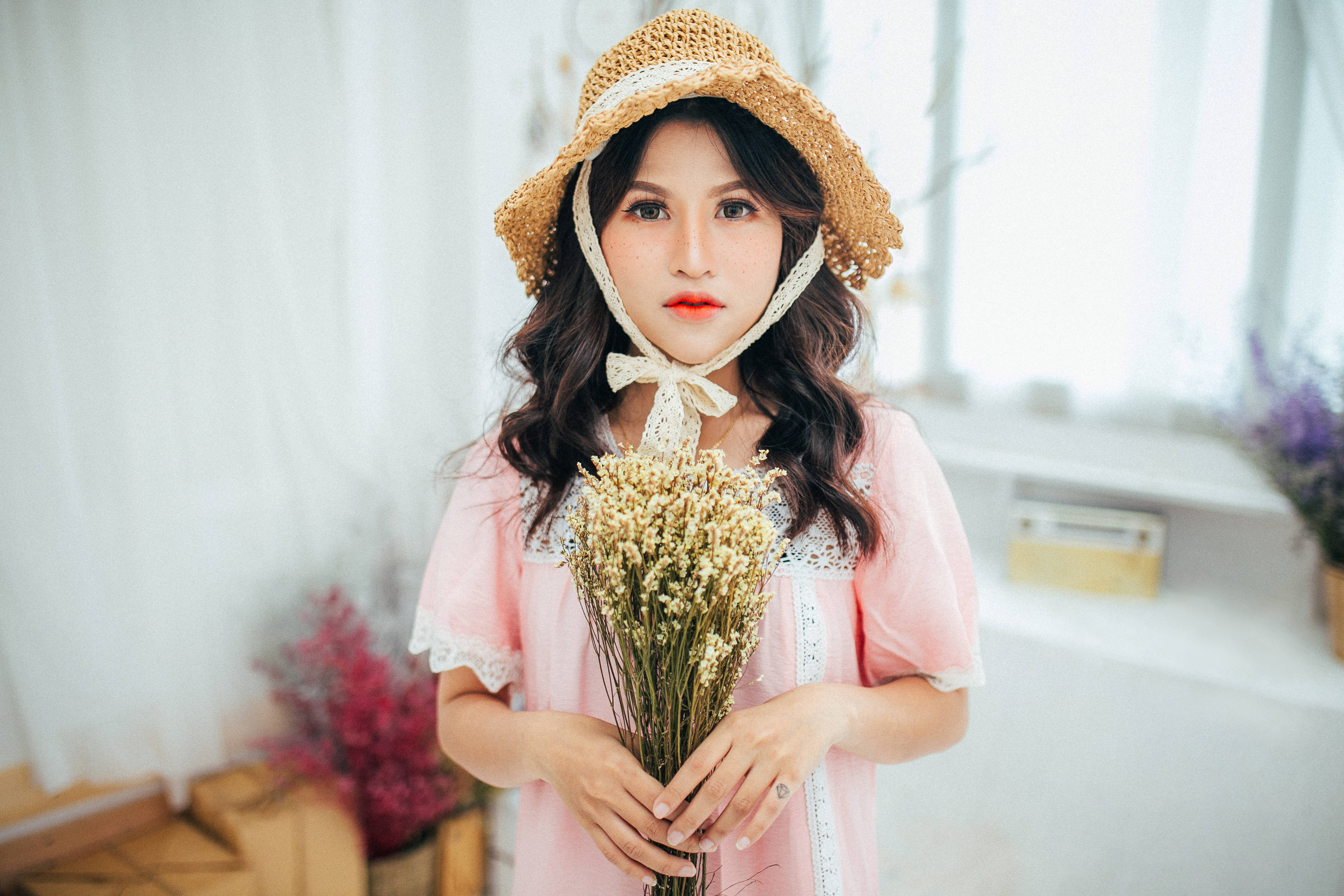 穿粉紅春季服裝的可愛女孩 壁紙 — 一位身穿粉紅色蕾絲連衣裙和草帽的年輕女性，在柔和光線的室內環境中手持乾花。